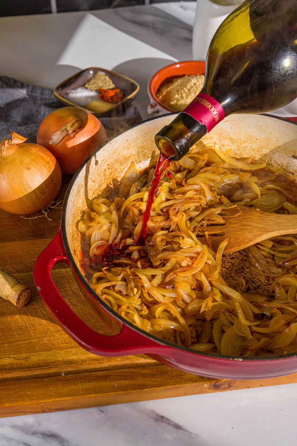 A cast iron skillet with caramelized onions and red wine deglazing the pan. The skillet is on a wooden board with onions.