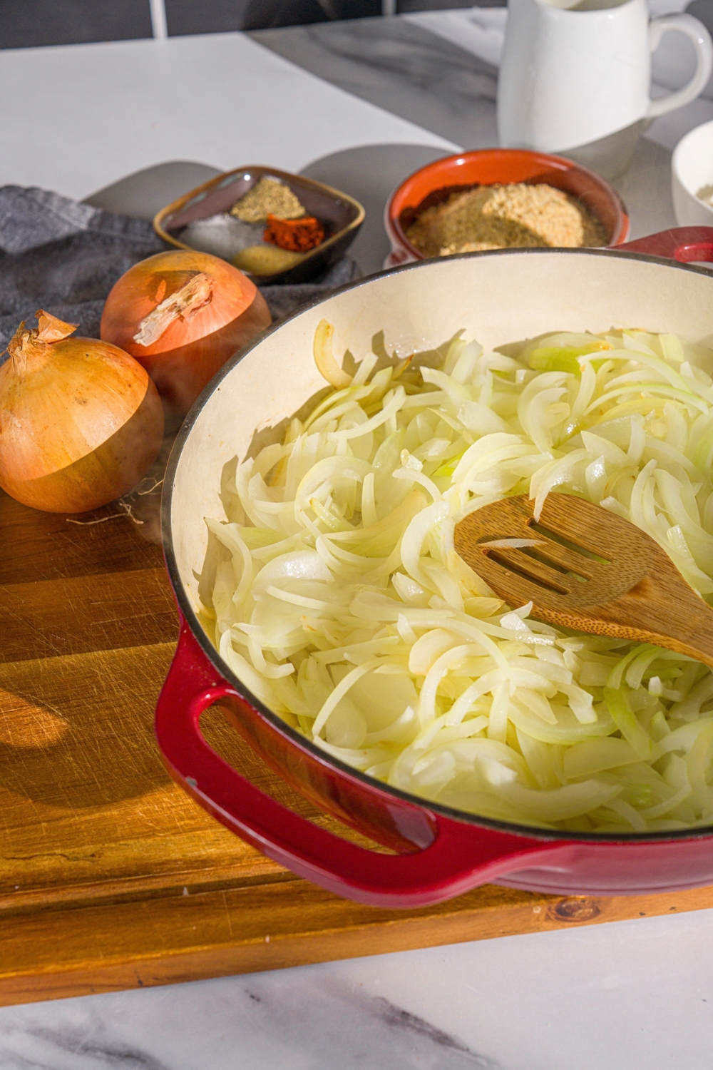 A cast iron skillet with sliced onions tossed in oil. There is a wooden spoon tossing the onions. The skillet is on a wooden board.