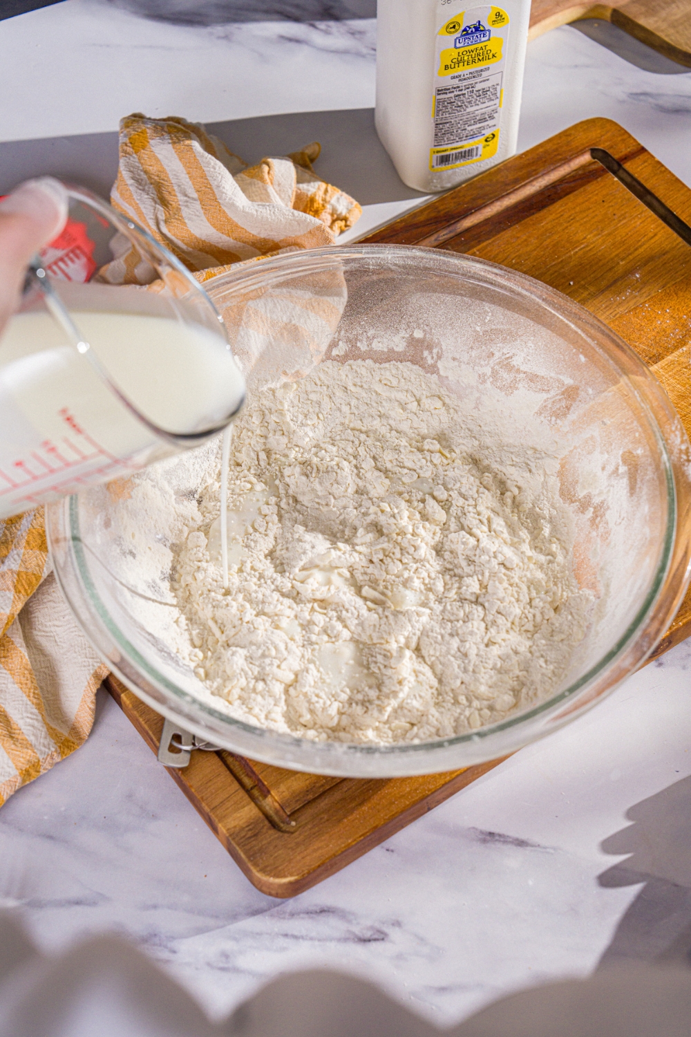 A large bowl of dry ingredient mixture with buttermilk being added to the bowl. The bowl is on a wooden board.