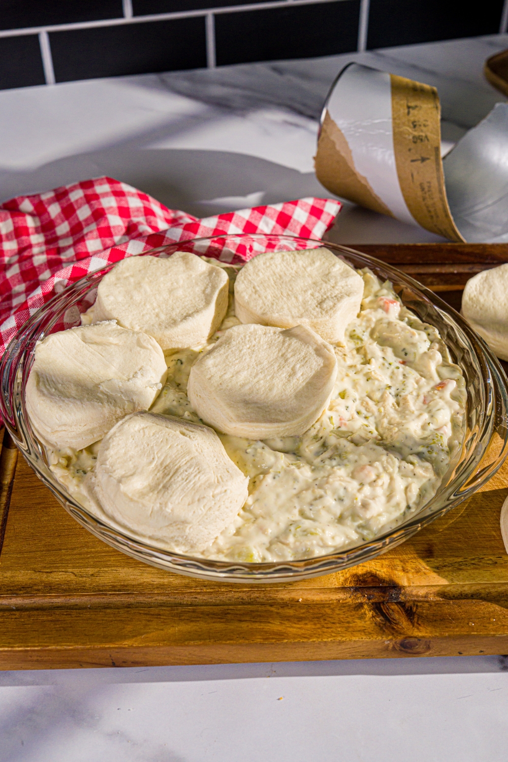 A round baking dish with chicken pot pie soup mixture topped with unbaked split biscuits. The dish is on a wooden board with a red checkered napkin.
