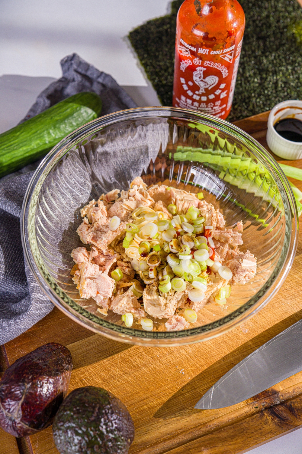 A glass bowl with ingredients to make tuna sushi mixture including tuna, kewpie mayo, sriracha, soy sauce, and sliced green onion. The bowl is on wooden board with ingredients.