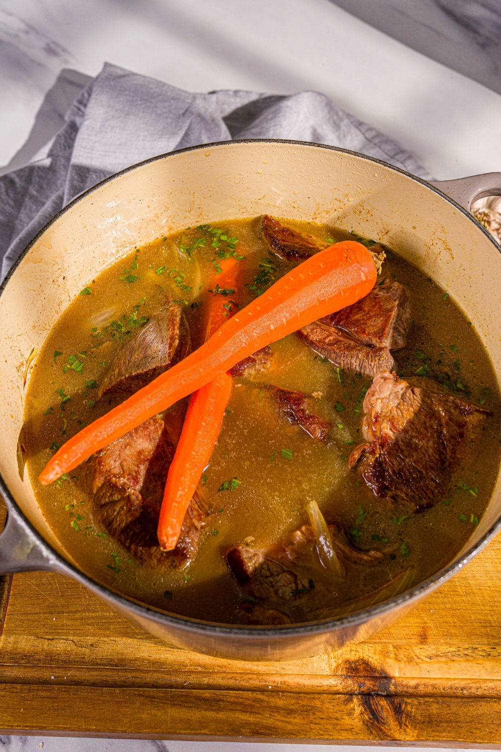 A dutch oven with browned beef chuck roast cooking in apple cider, carrots, and herbs. The dutch oven is on a wooden board.