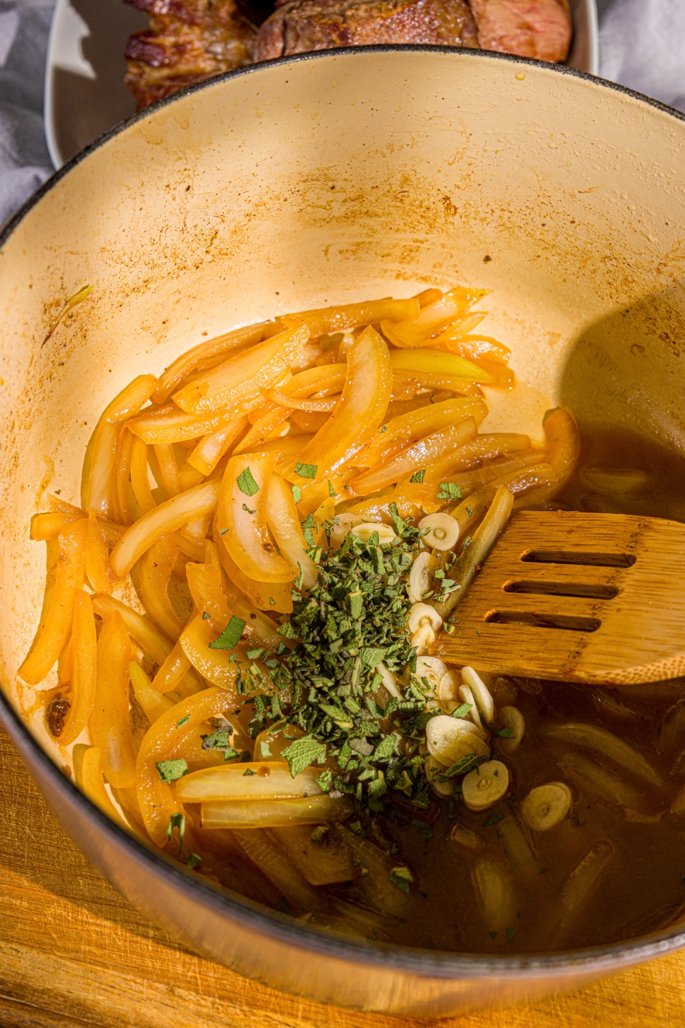 A dutch oven with sautéed sliced onion, garlic, and herbs deglazing in the pot. The dutch oven is on a wooden board.