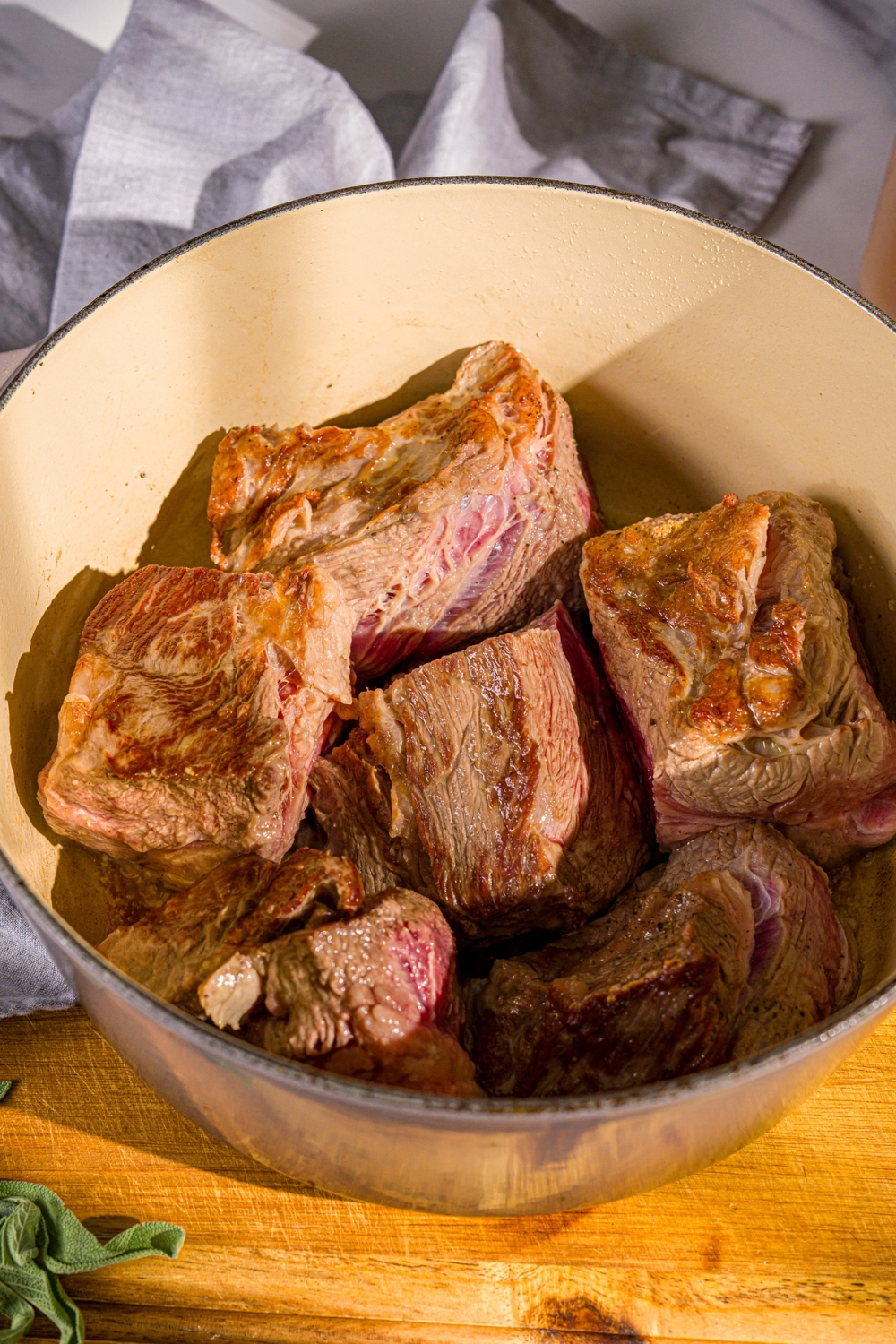 A dutch oven with browned chuck roast pieces. The dutch oven is on a wooden board.