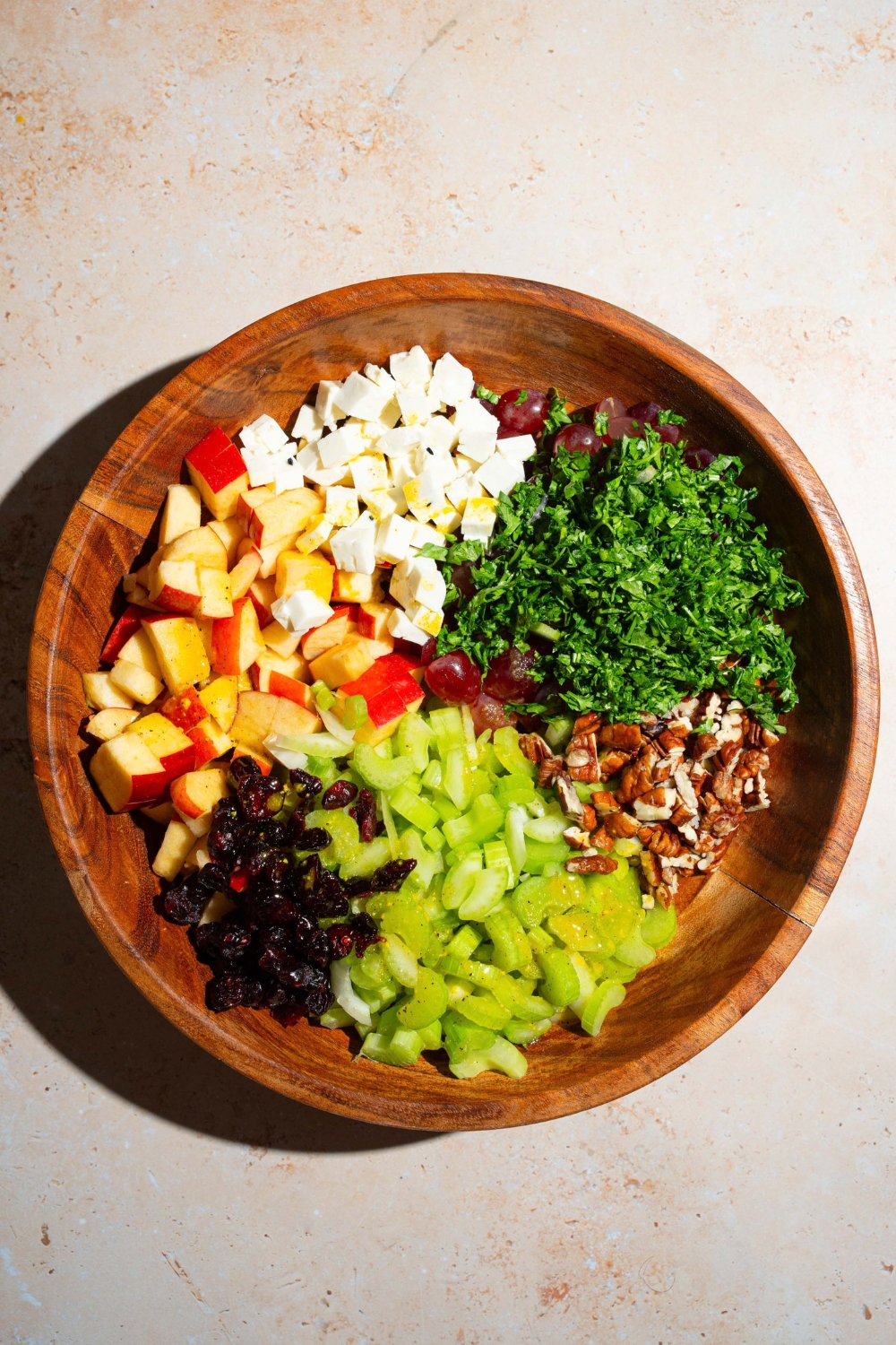 A wooden bowl with ingredients to make apple crunch salad including apples, grapes, celery, feta cheese, dried cranberries, pecans, and feta cheese. The bowl is on a tan counter.