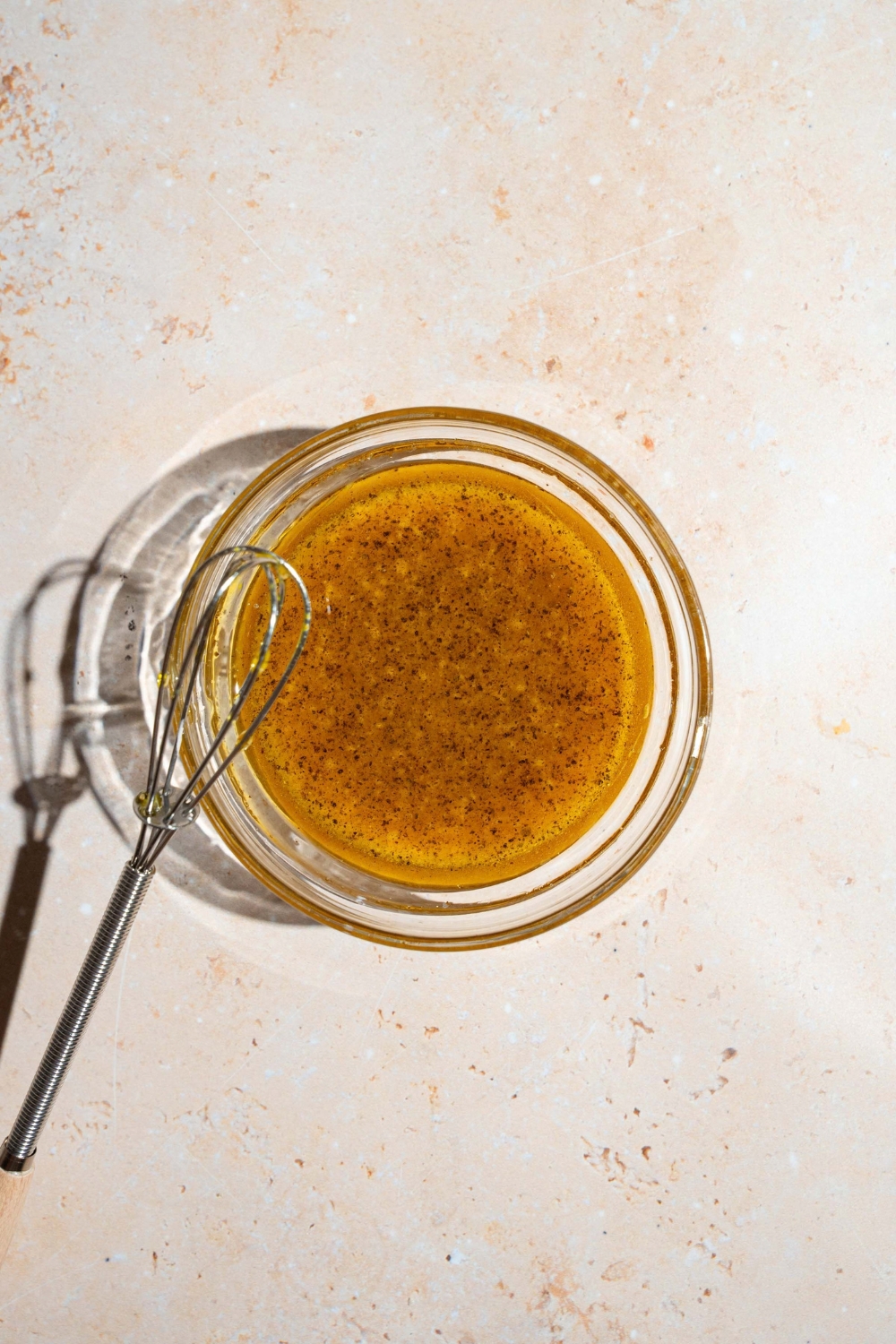 A small glass bowl with a maple dijon dressing mixture. There is a whisk on the bowl. The bowl is on a tan counter.