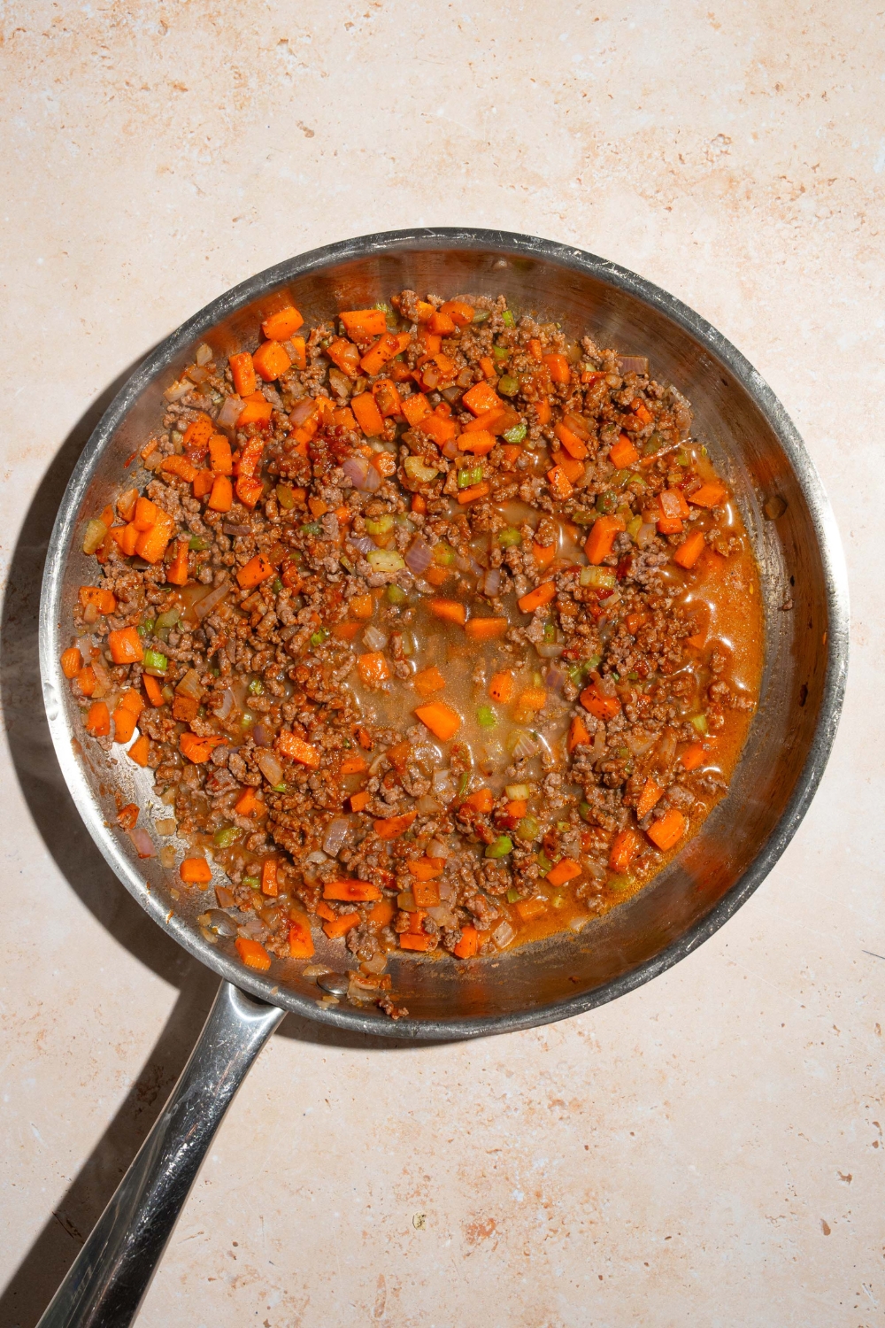 A skillet with a browned beef and sautéed vegetable mixture with broth and seasonings. The skillet is on a tan counter.