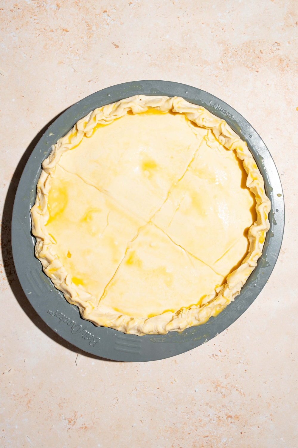 A whole unbaked meat pot pie on a gray platter. The top of the pie is covered with egg wash. The platter is on a tan counter.