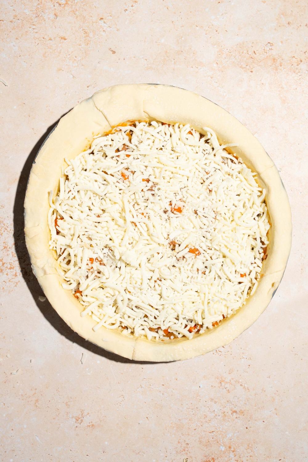 A pie plate shaped with pie crust and filled with beef pot pie filling, and topped with shredded mozzarella. The plate is on a tan counter.
