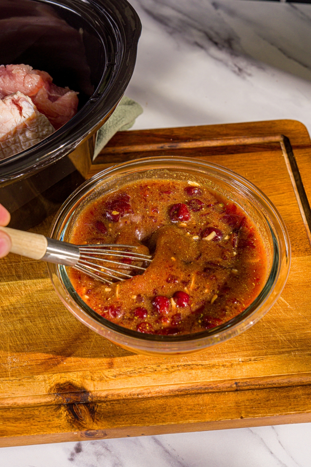 A small bowl with a whisk mixing ingredients to make cranberry sauce for crock pot butterball turkey breast including orange juice, apple cider, and cranberry sauce. The bowl is on a wooden board.