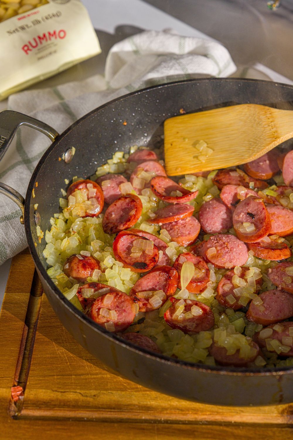 A skillet with sautéed onions and sliced sausages. There is a wooden spoon stirring the mixture. The skillet is on a wooden board.