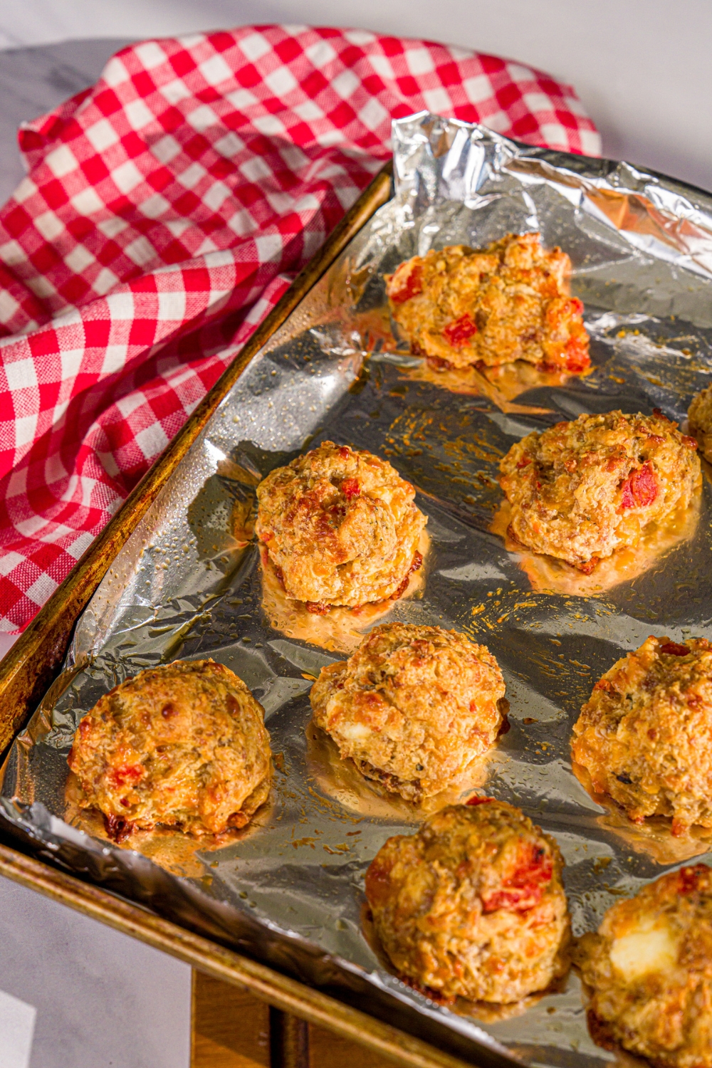 A baking sheet lined with foil with baked cheddar bay biscuit sausage balls. The sheet is on a wooden board with a red checkered napkin.