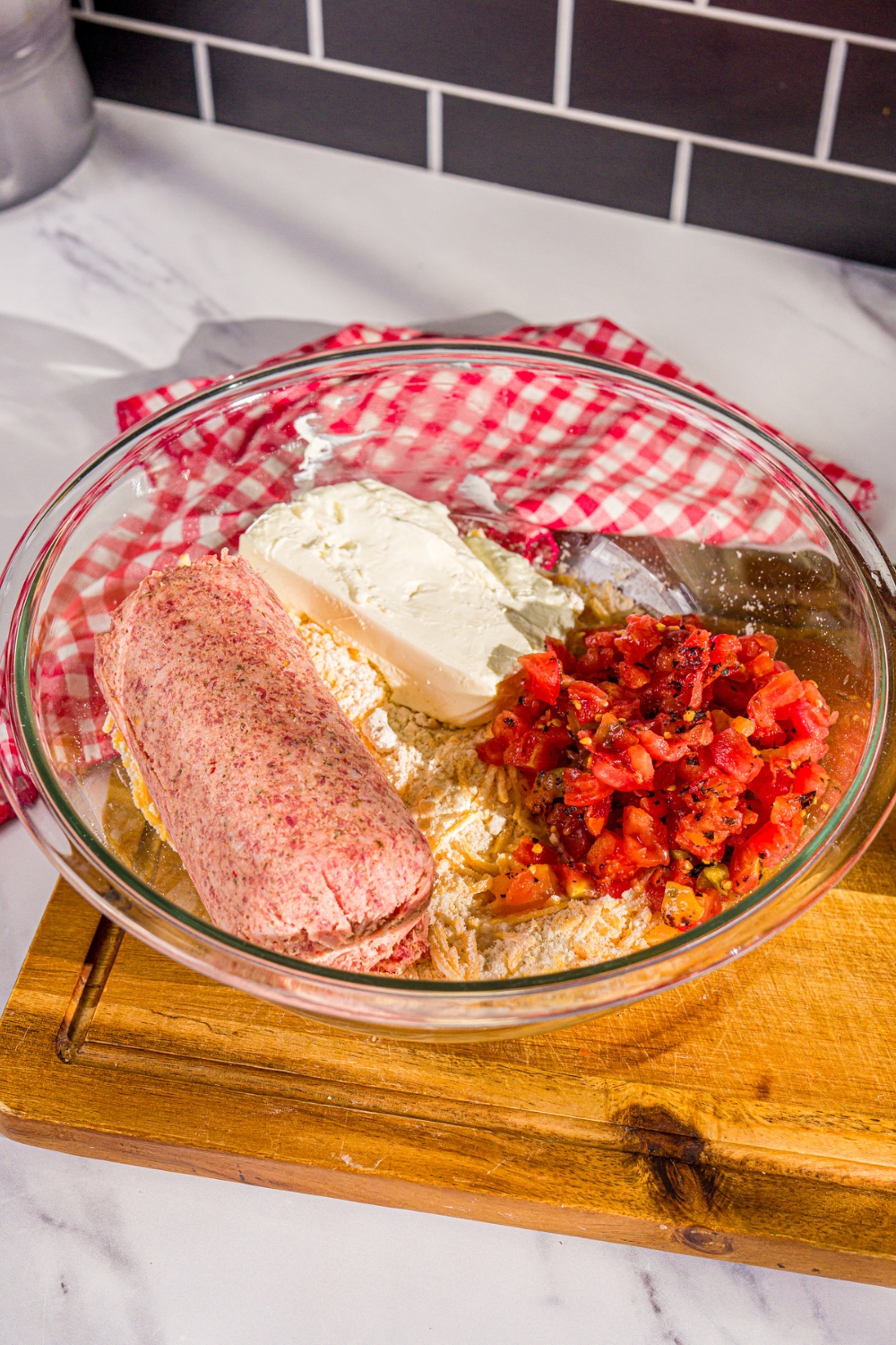 A large bowl with a biscuit mix and sausage mixture including cream cheese, biscuit mix, shredded cheese, rotel, and sausage. The bowl is on a wooden board with a red checkered napkin.
