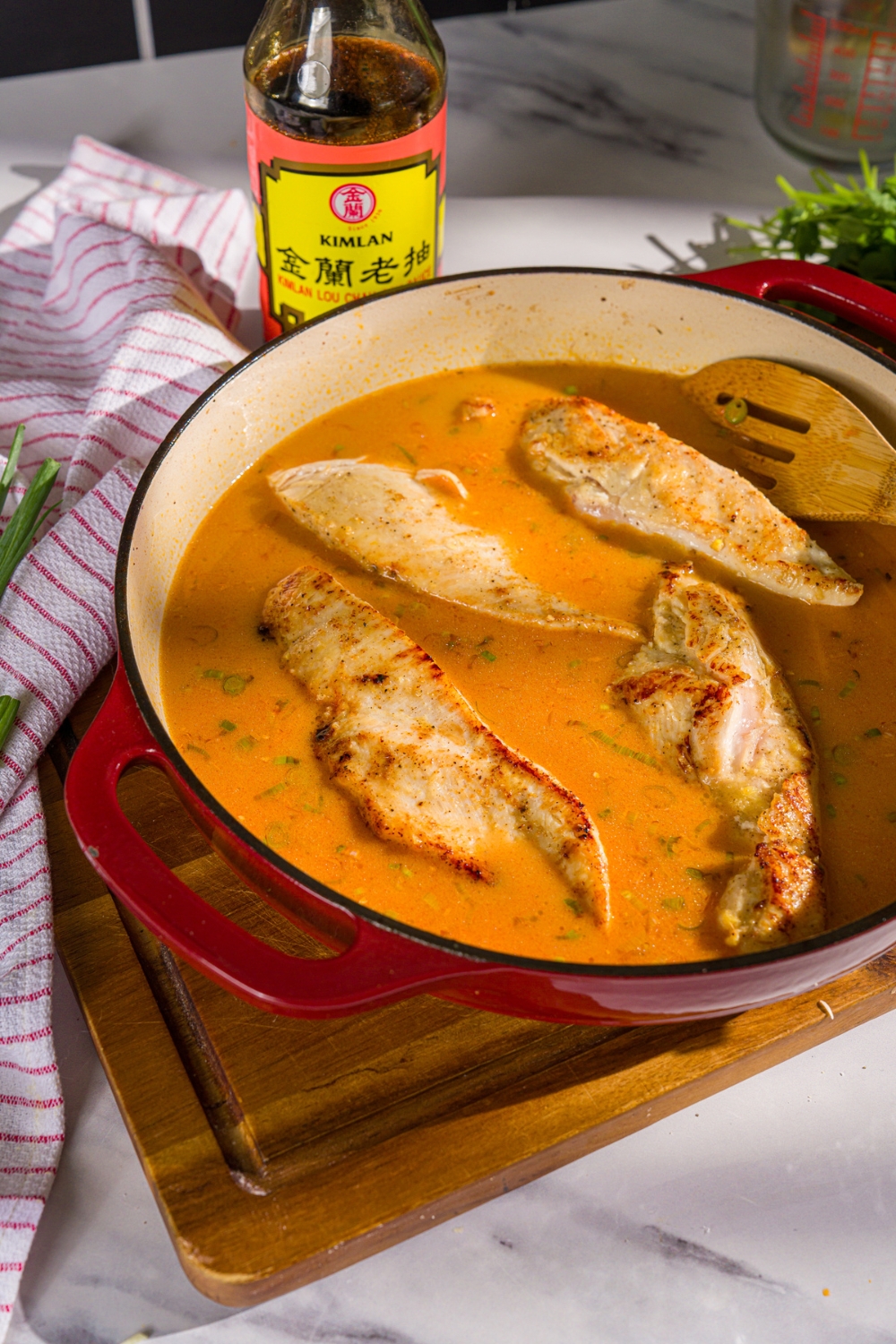 A skillet with seared chicken breasts in a thai coconut broth. The skillet is on a wooden board with a white striped napkin.