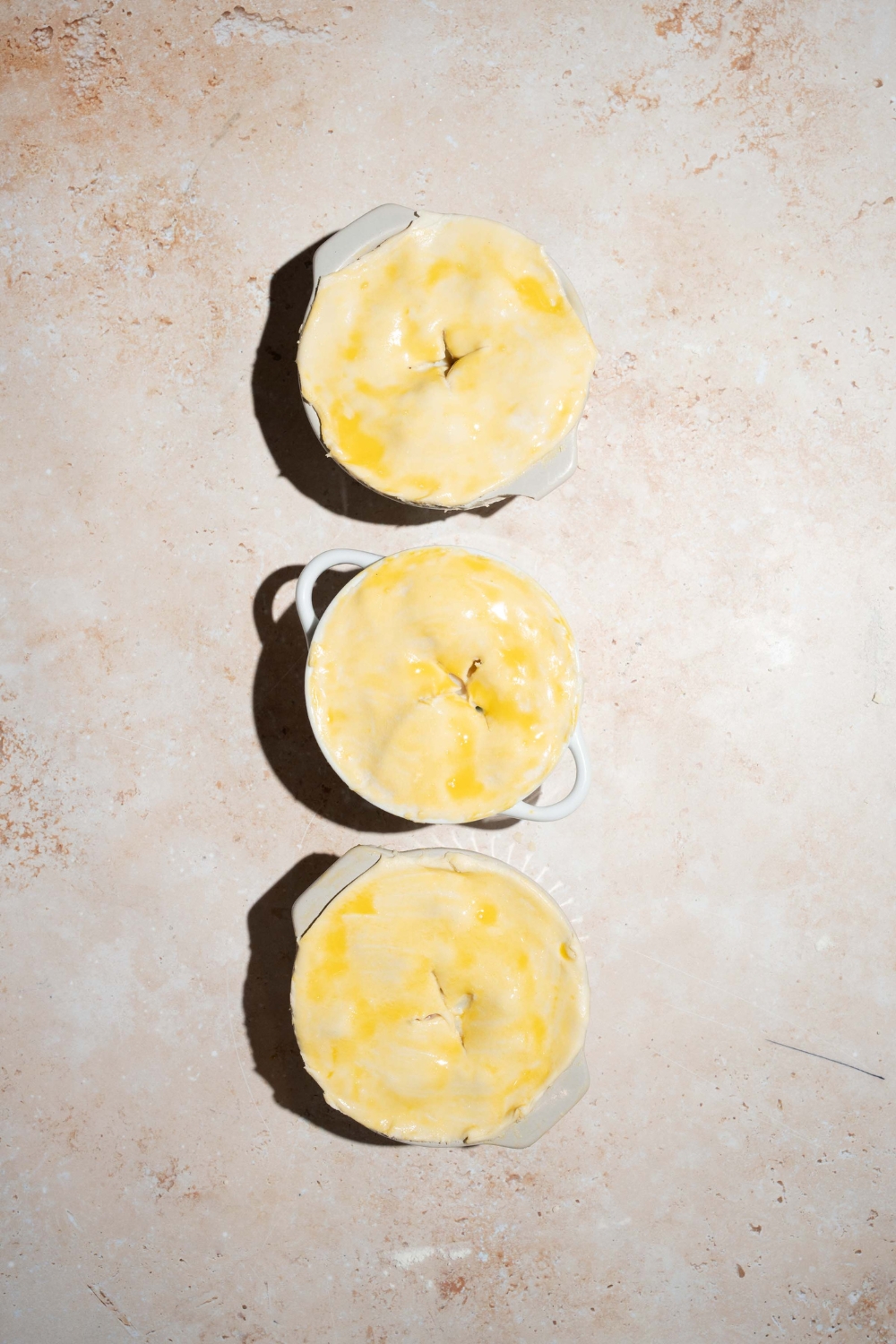 Three small pie tins filled with KFC pot pie filling and topped with uncooked puff pastry. The tins are on a tan counter.