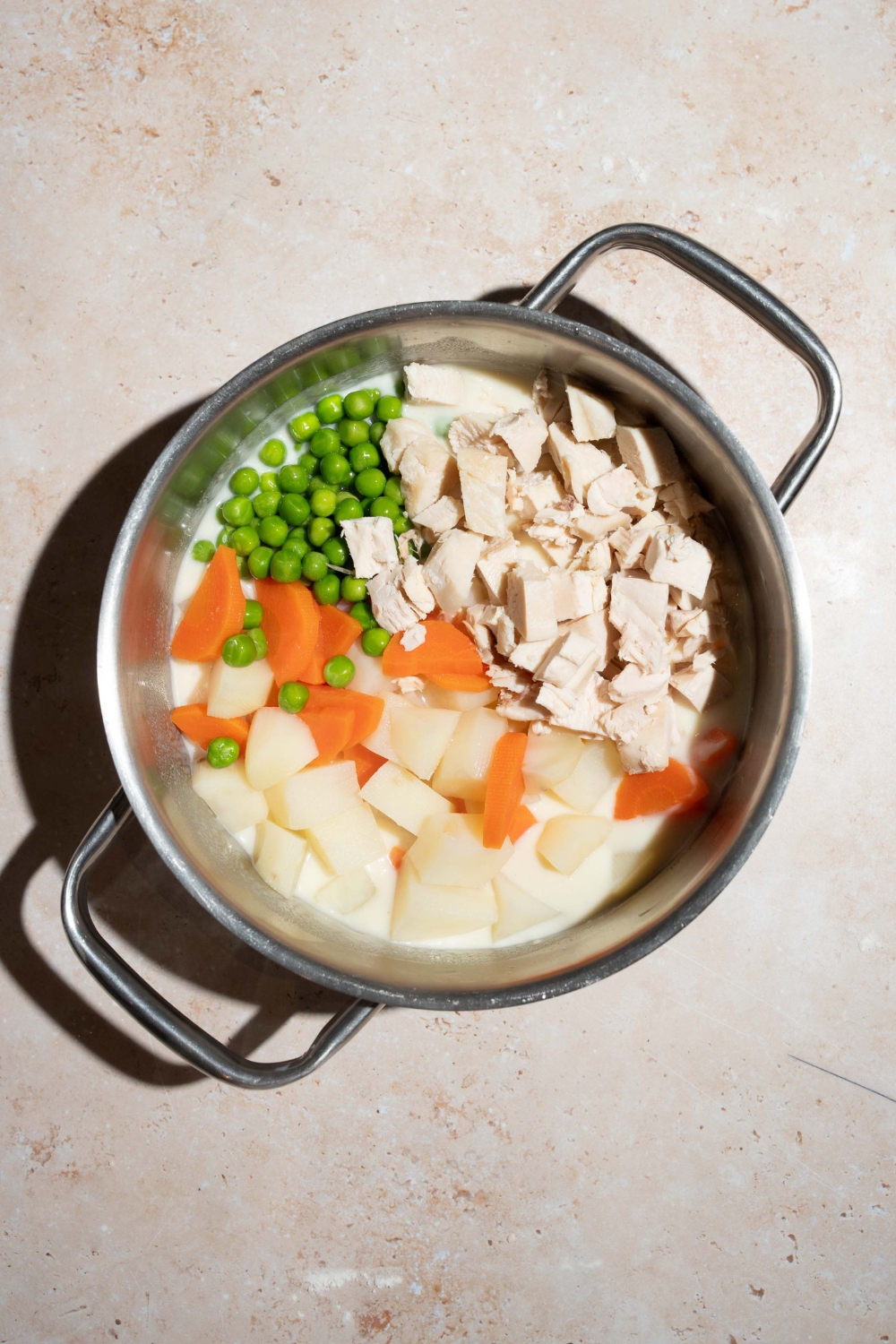 A large pot with pot pie filling ingredients including chicken, potatoes, carrots, and peas in a milk and soup mixture. The pot is on a tan counter.