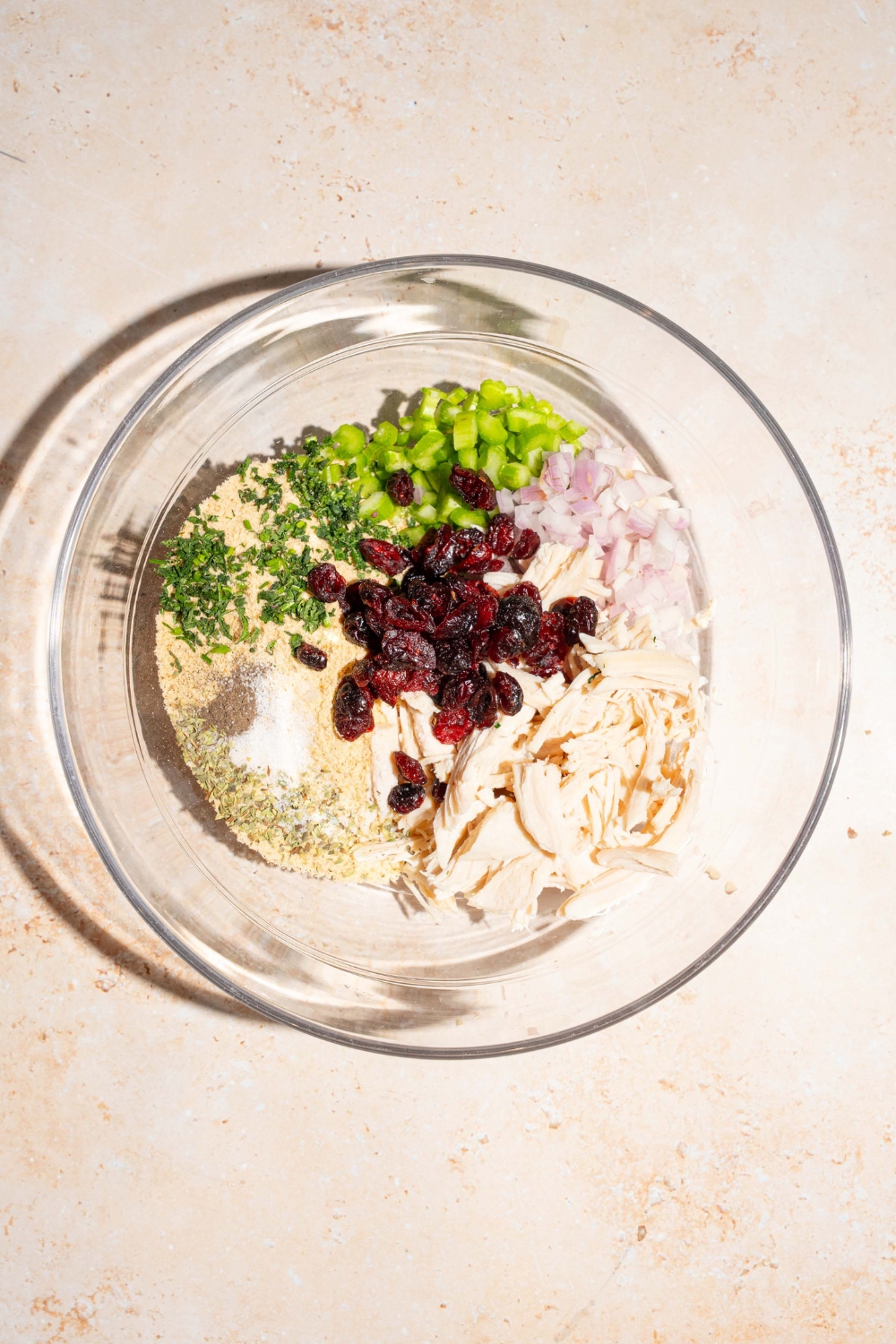 A large bowl with ingredients to make cranberry turkey stuffing mixture including turkey, dried cranberries, celery, onion, and breadcrumbs. The bowl is on a tan counter.