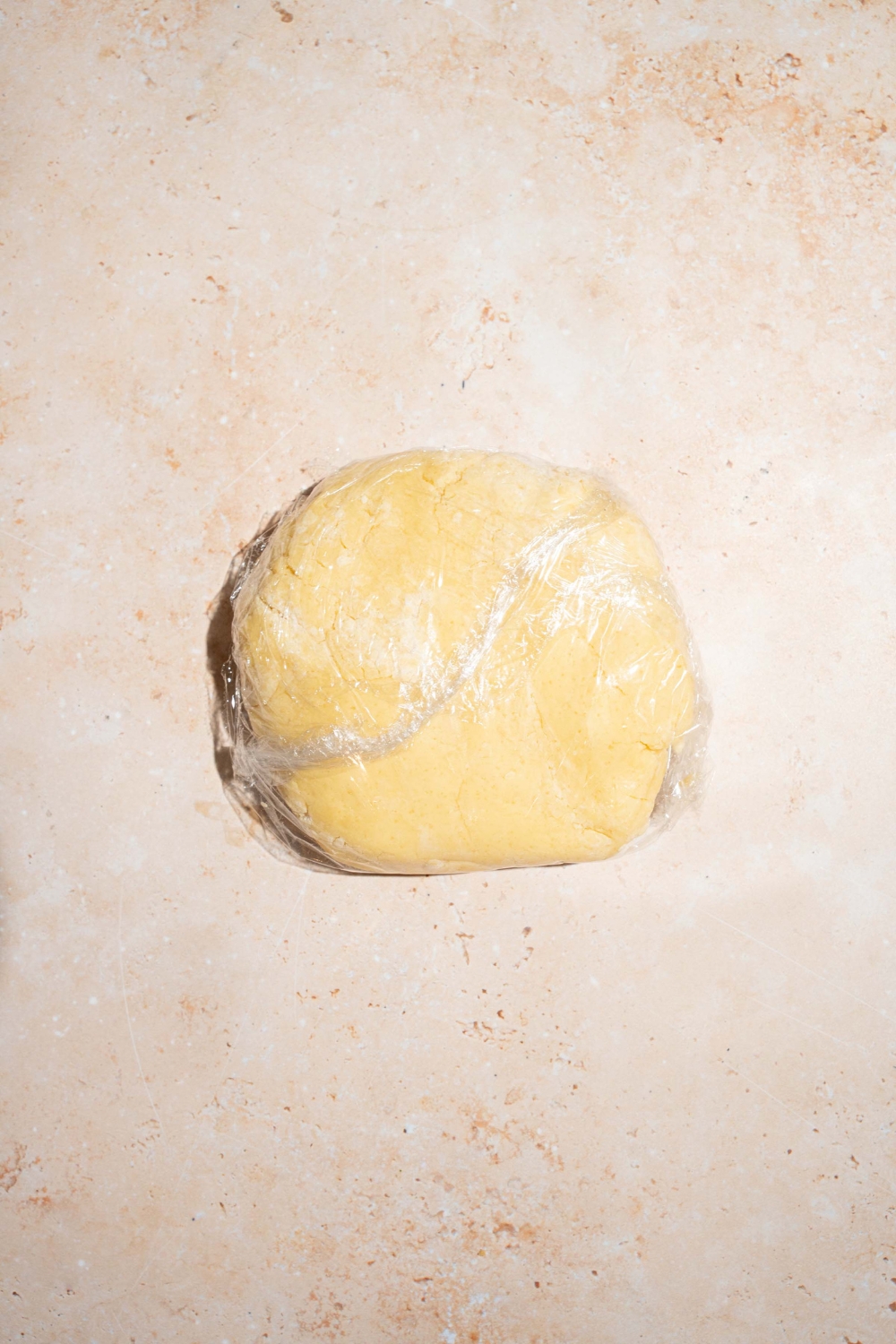 A ball of pie crust dough wrapped in saran wrap on a tan counter.