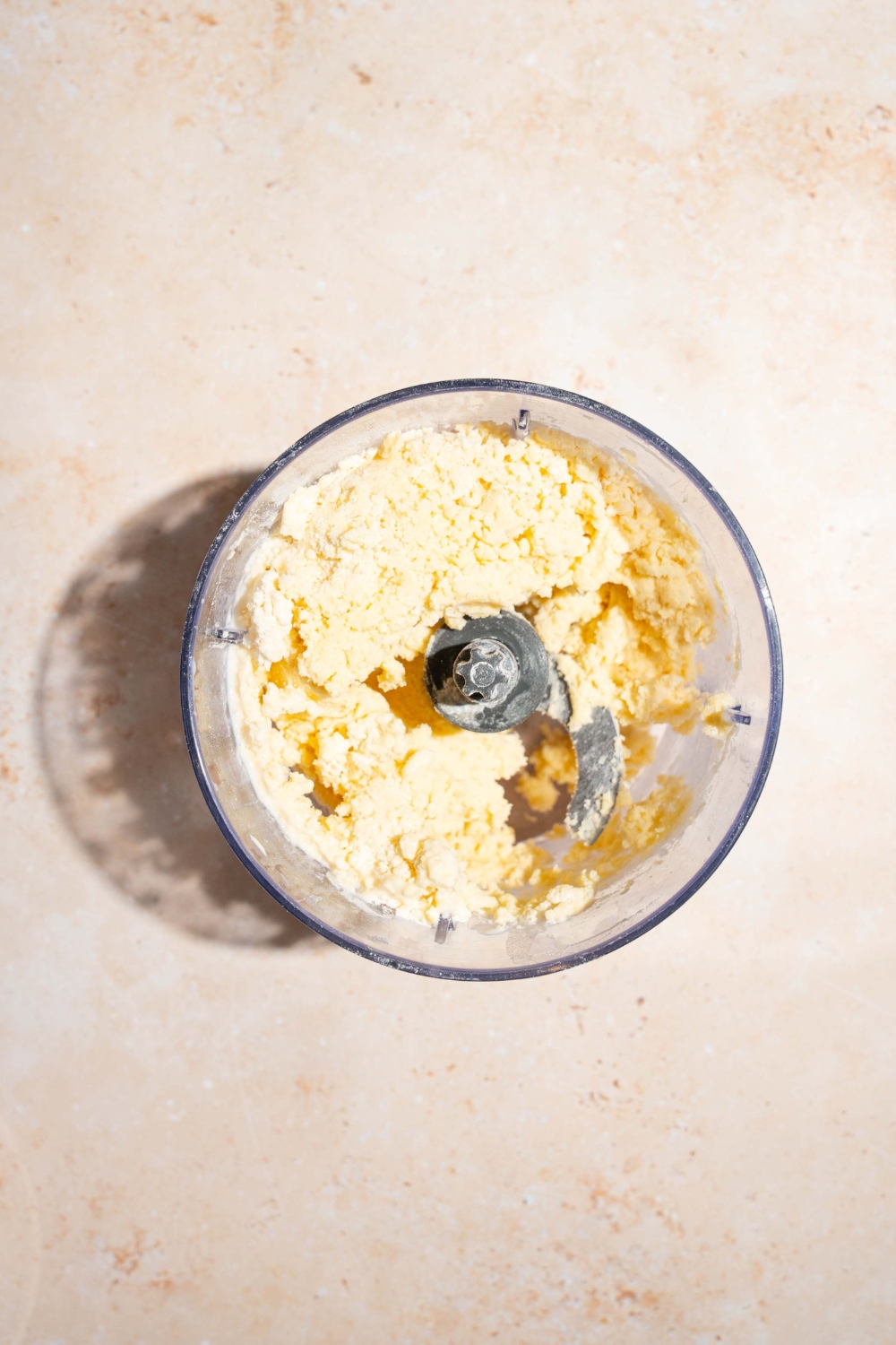 A food processor with pulsed pie crust dough ingredients including flour, sugar, and butter. The food processor is on a tan counter.