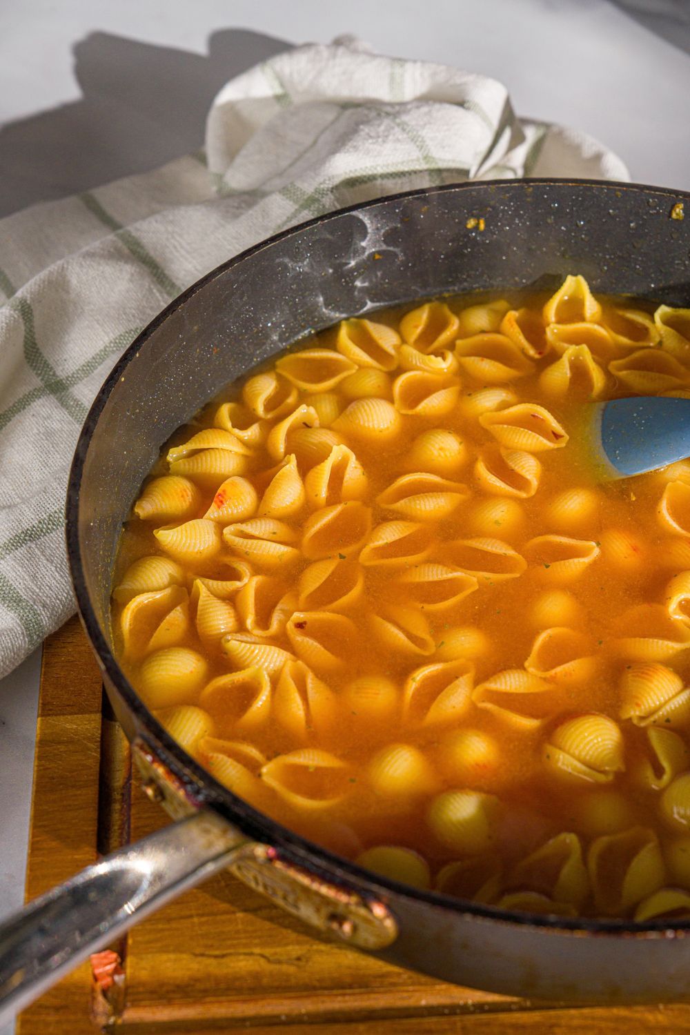 A skillet with shell pasta in chicken broth and tomato paste mixture. The skillet is on a wooden board.