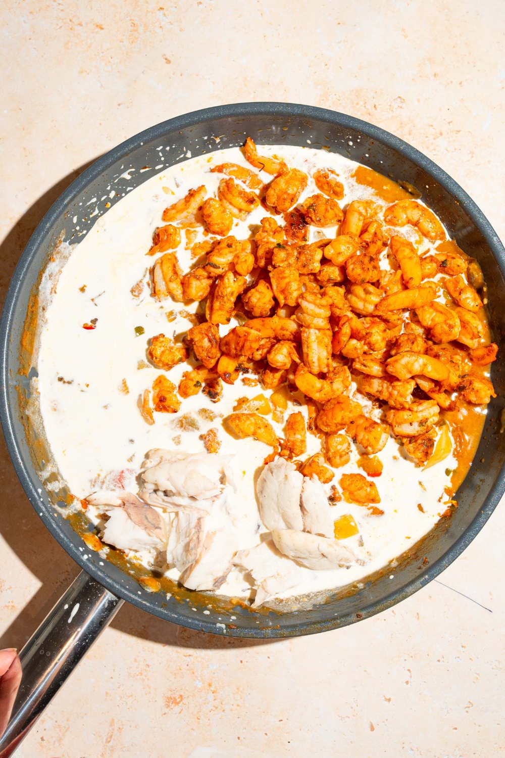 A skillet with browned seasoned shrimp with sautéed vegetables and broth with cream being added to the mixture. The skillet is on a tan counter.