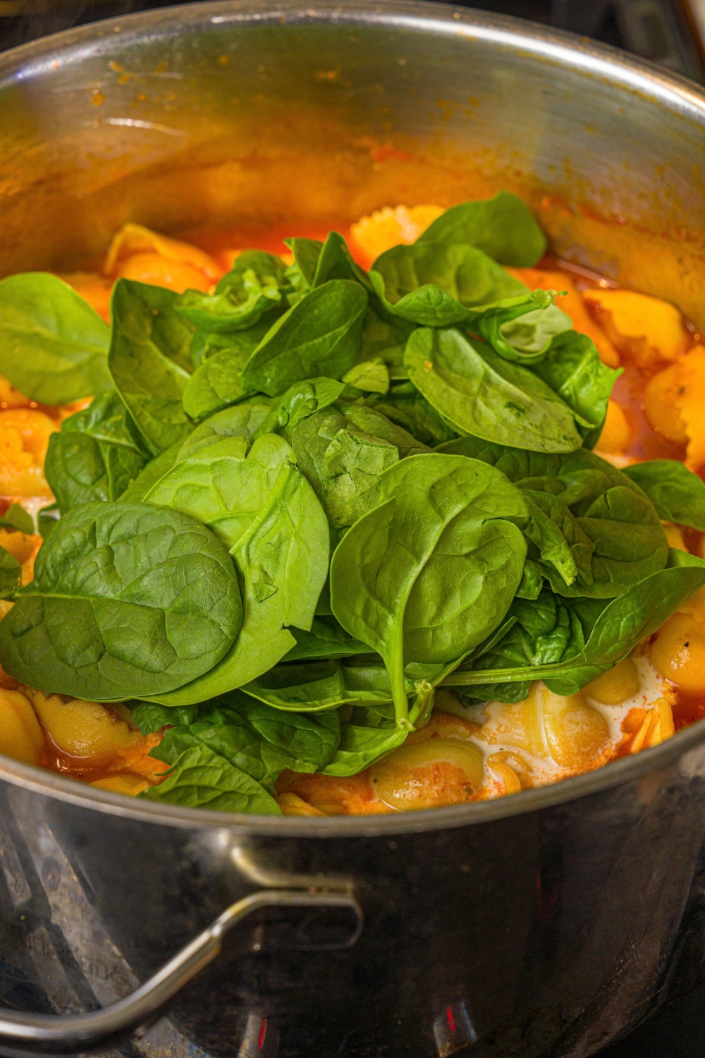 A large pot with a tomato tortellini soup mixture topped with spinach. The pot is cooking on the stove.