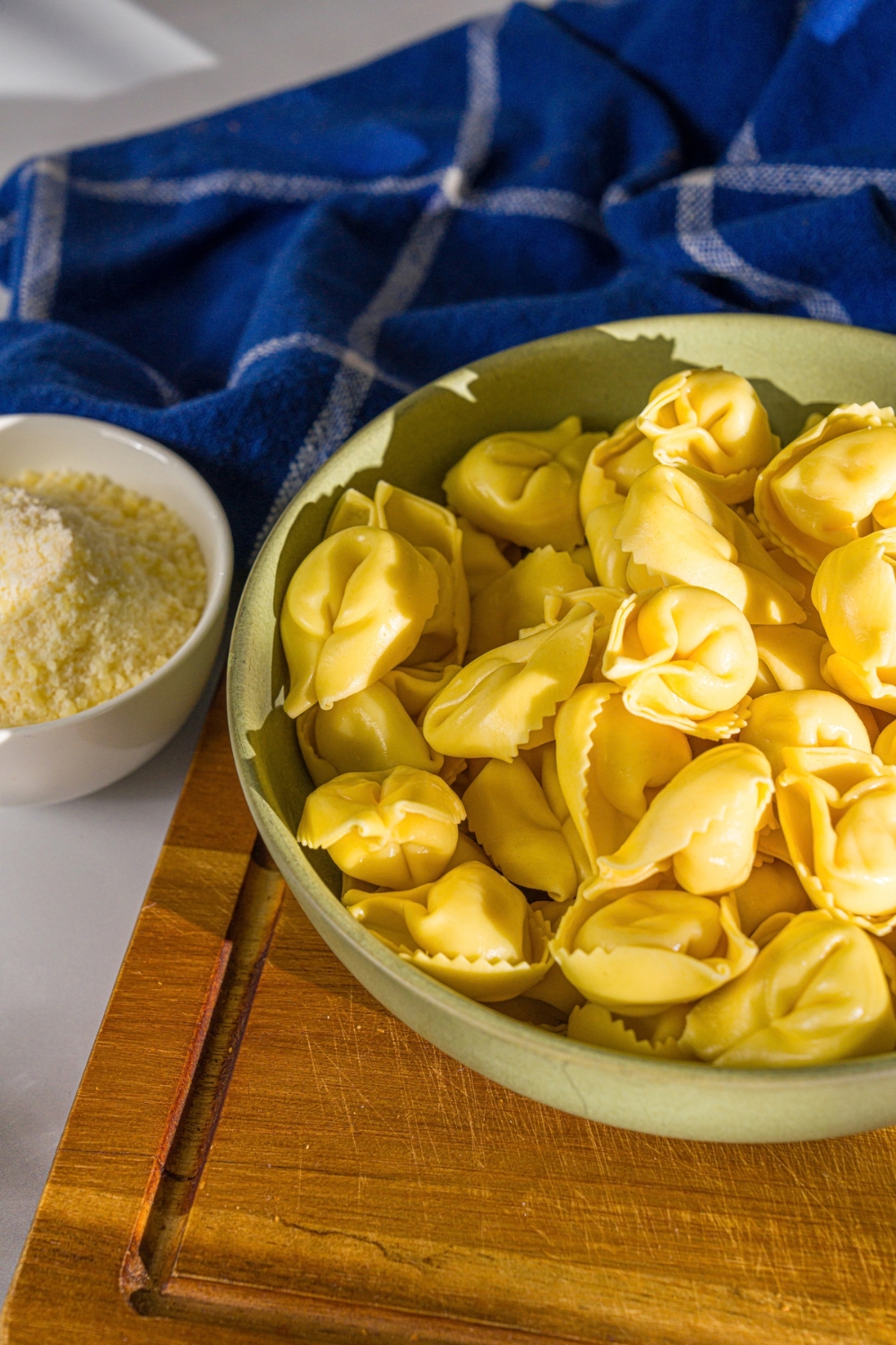 A bowl of cooked tortellini pasta. The bowl is on a wooden board with a small bowl of cheese and a blue cloth napkin.
