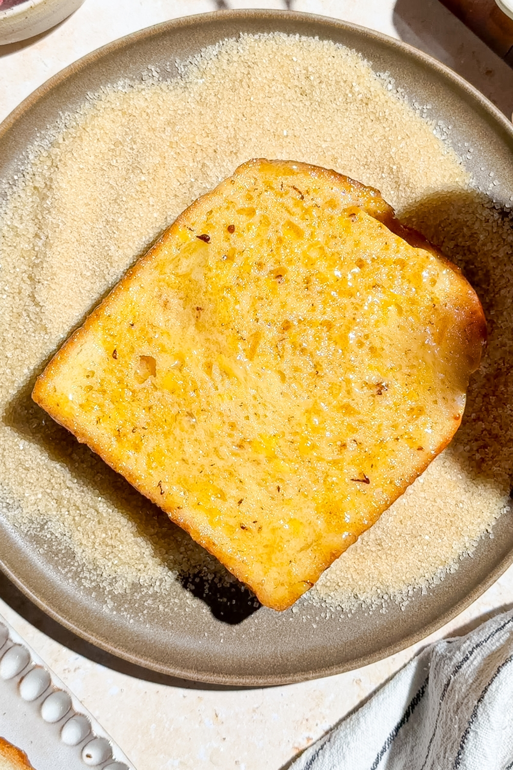 A slice of soaked brioche bread in a plate of turbinado sugar. The plate is on a tan counter.