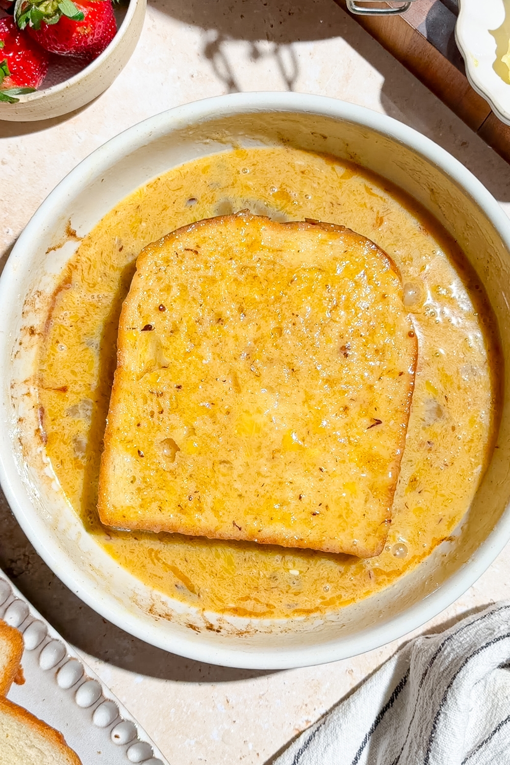 A slice of brioche bread soaking in an egg and milk mixture. The bowl is on a tan counter with a bowl of strawberries.