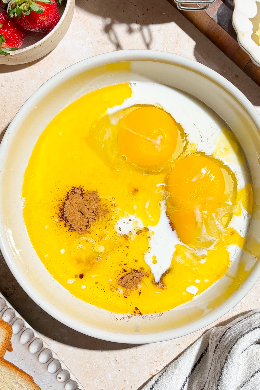 A bowl with eggs, milk, cinnamon, butter, and brown sugar. The bowl is on a tan counter.
