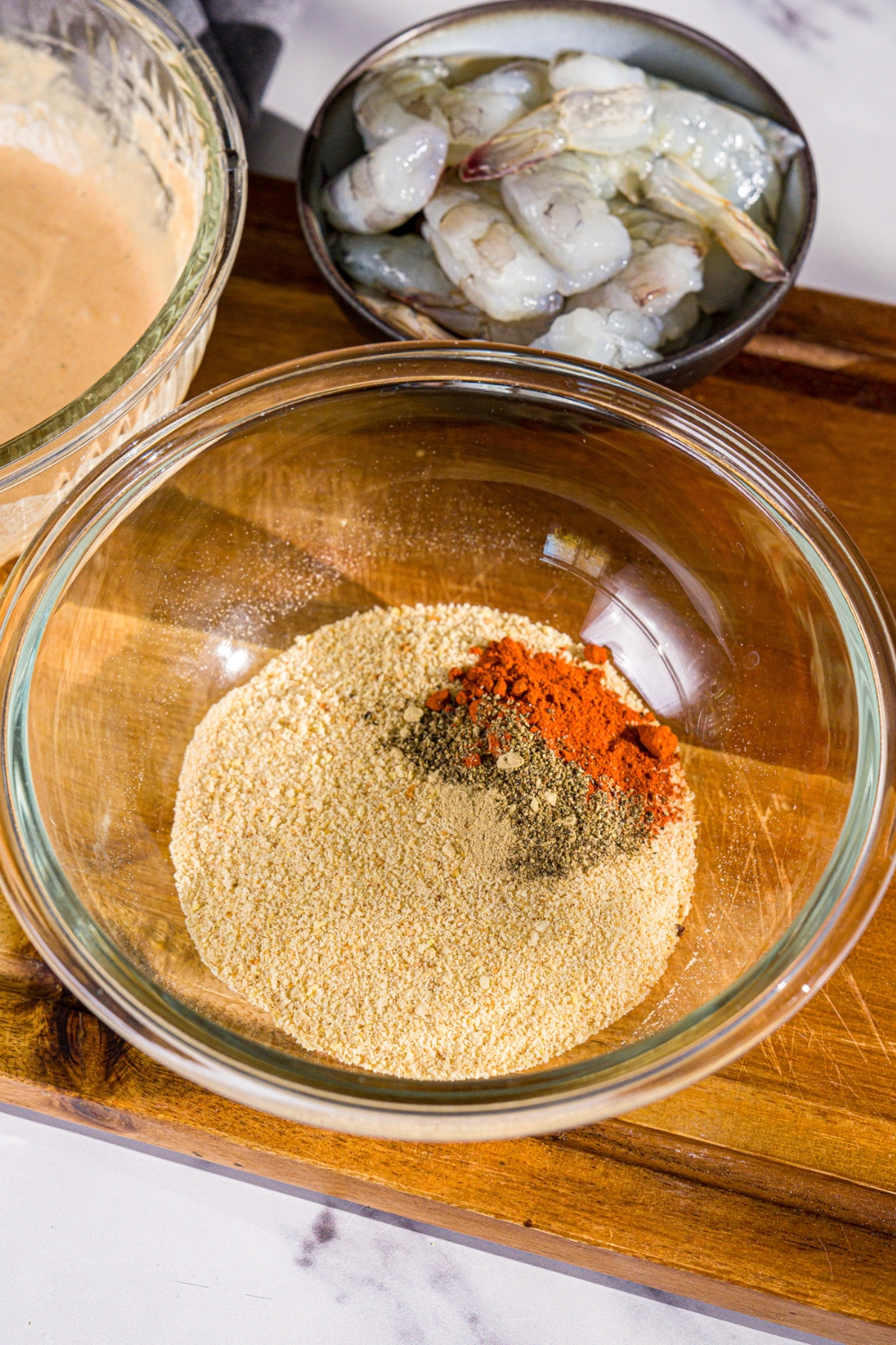 A bowl with ingredients to make fried prawn breading including breadcrumbs, peri peri seasoning, pepper, and paprika. The bowl is on a wooden board with a bowl of prawns and a bowl of batter.