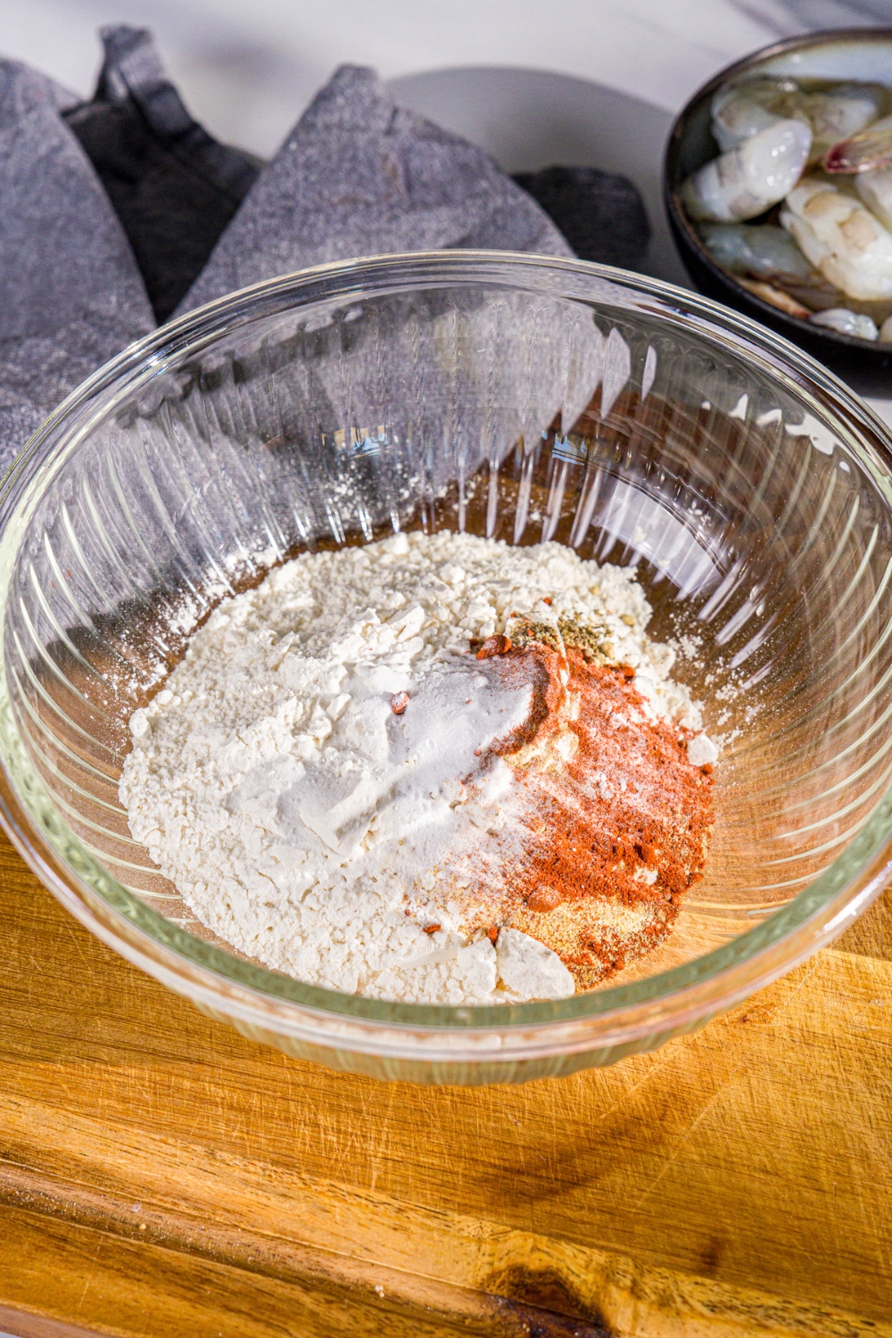 A bowl of ingredients to make crispy fried prawn batter including flour, pepper, peri peri seasoning, garlic powder, and paprika. The bowl is on a wooden board with a blue cloth napkin.