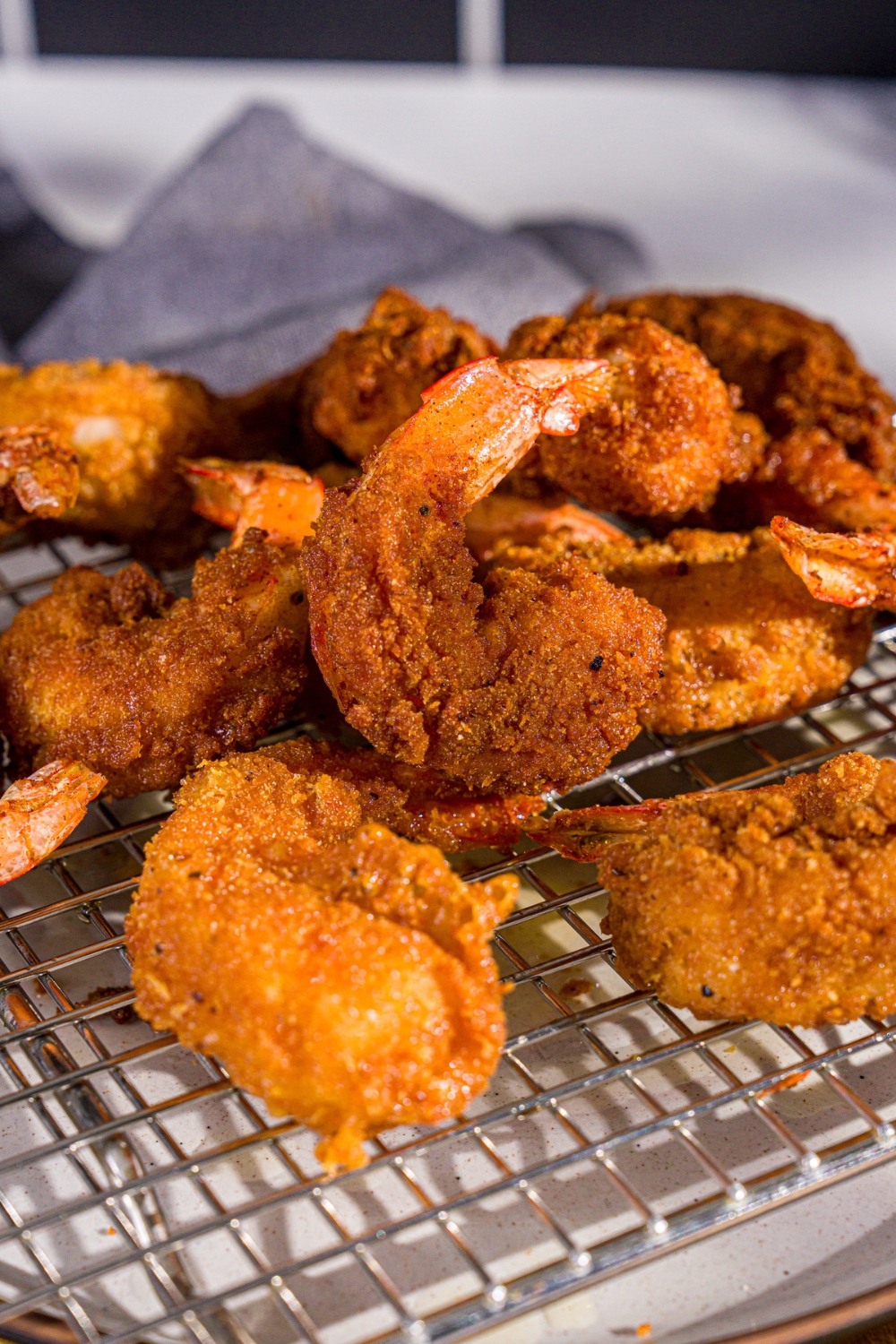Crispy fried prawns on a drying rack over a white plate. The plate is on a wooden board with a blue cloth napkin.