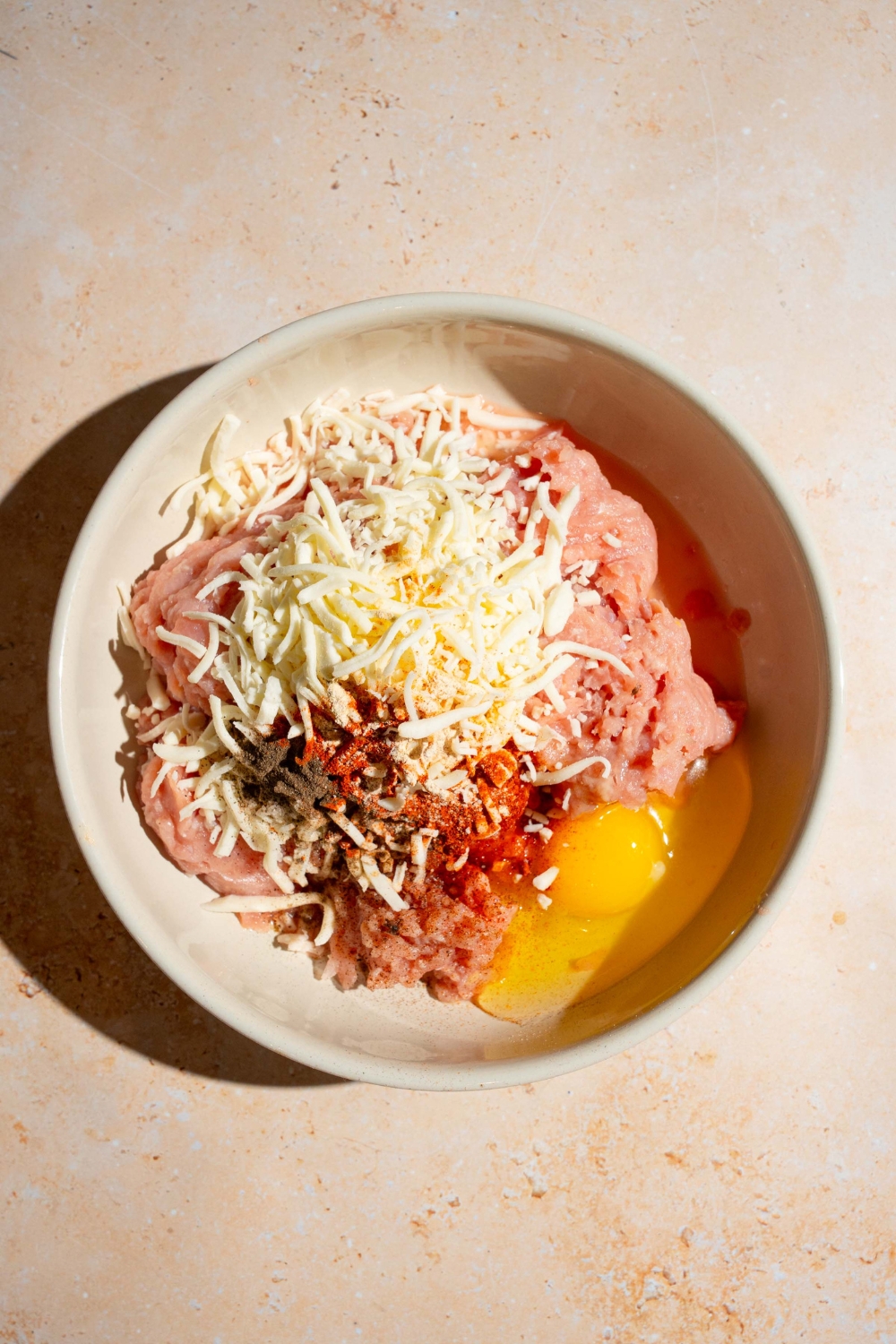 A bowl of ingredients to make chicken filling including ground chicken, egg, shredded mozzarella, and seasonings. The bowl is on a tan counter.