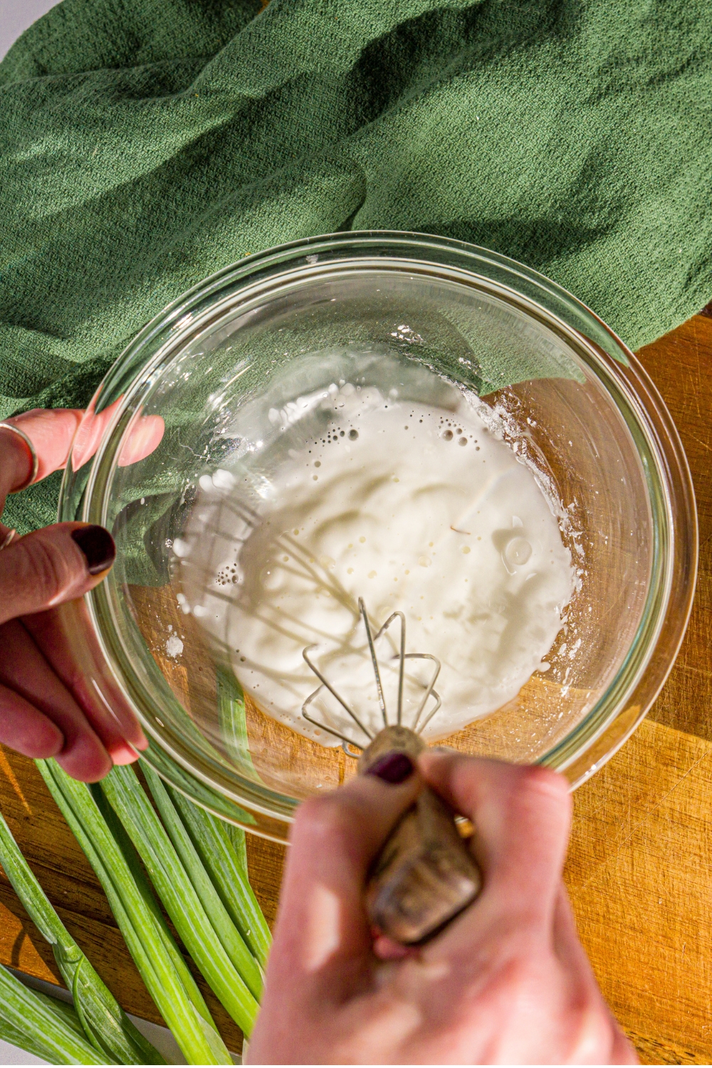 A small bowl with a whisk mixing a slurry with cornstarch and water. The bowl is on a wooden board with green onions and a green cloth napkin.