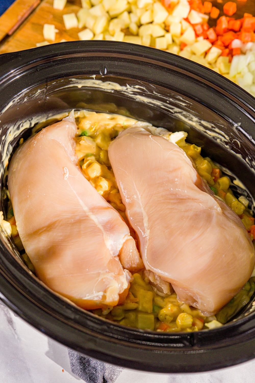 A crockpot with a vegetable and soup pot pie mixture topped with two uncooked chicken breasts. The pot is on a marble counter.