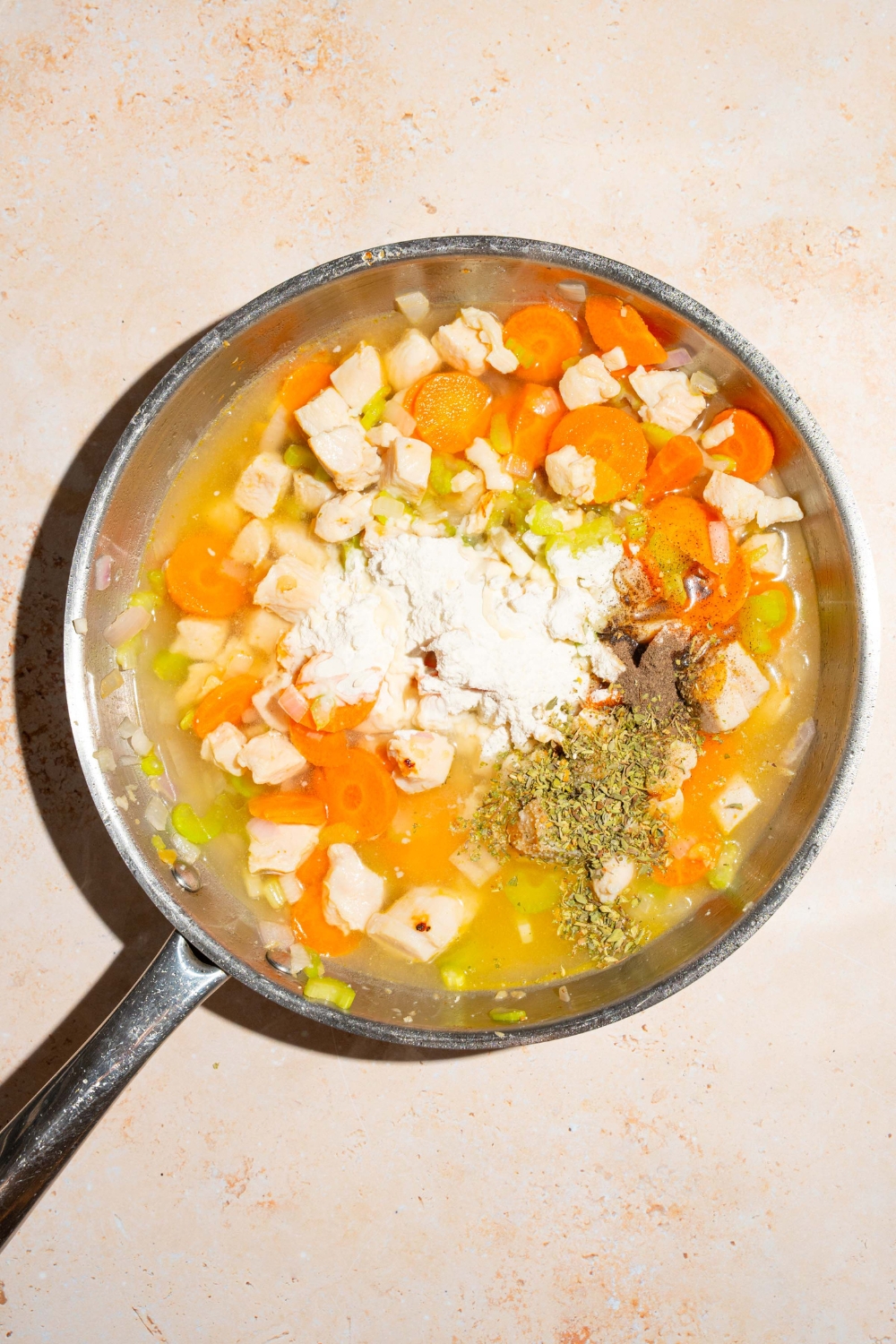 A skillet with sautéed chicken and vegetables in broth topped with flour and seasonings. The skillet is on a tan counter.