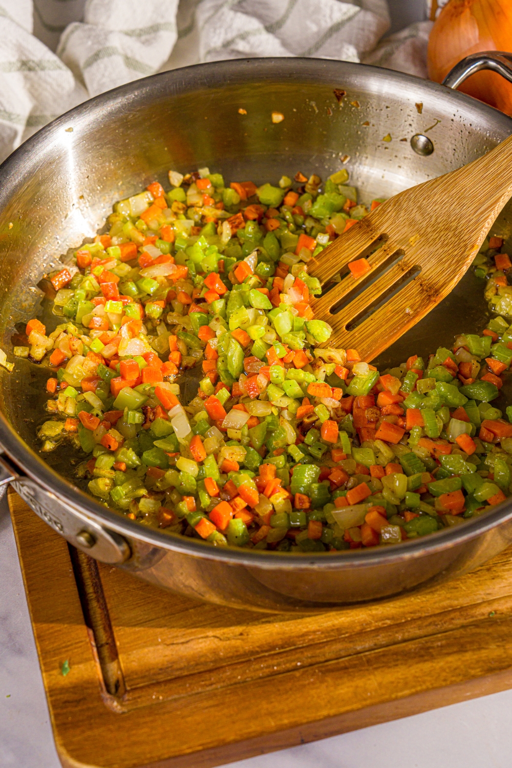A large skillet with chopped celery, carrot, and onion sautéing in oil. The skillet is on a wooden board.