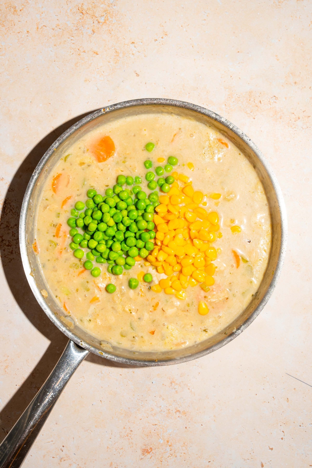 A skillet with chicken and vegetables in a broth mixture topped with peas and corn. The skillet is on a tan counter.