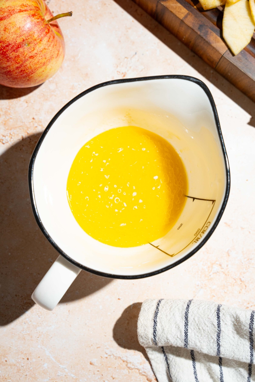 A large measuring pitcher with a mixture of wet ingredients to make invisible apple cake. The bowl is on a tan counter with a cutting board of sliced apples.