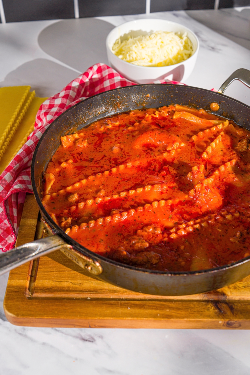 A skillet with one pan lasagna noodles cooked in tomato sauce. The skillet is on a wooden board with a red checkered napkin and a bowl of shredded cheese.