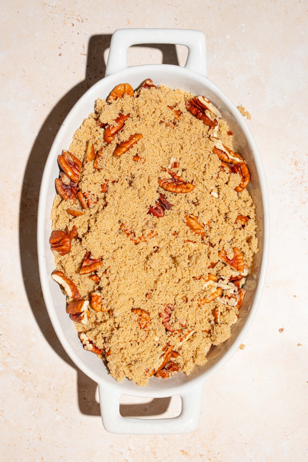 A casserole dish with a pecan pie cobbler batter topped with brown sugar and pecans. The dish is on tan counter.