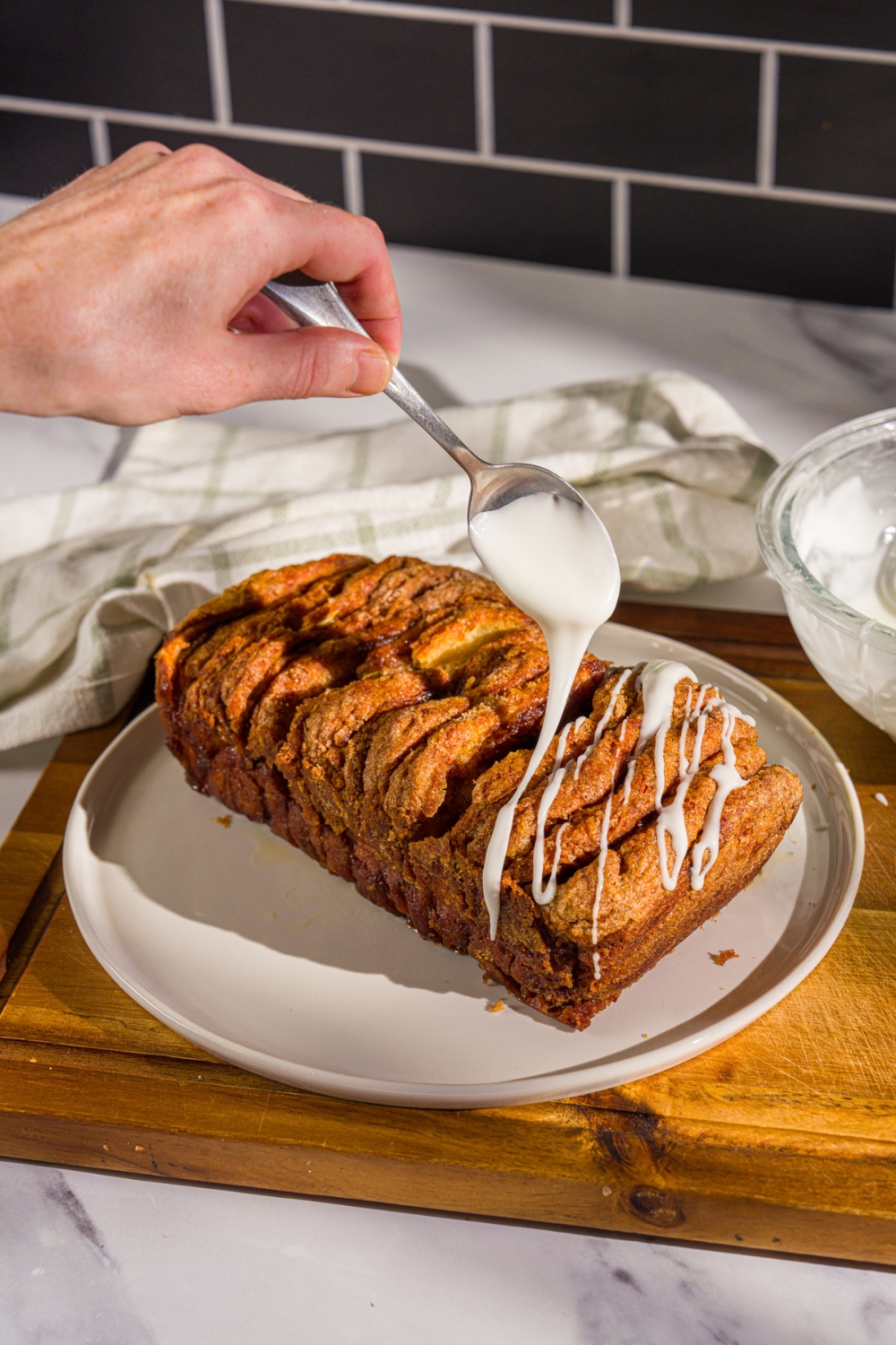 A white plate with a loaf of pumpkin pull apart bread. A spoon is drizzling glaze over the loaf. The plate is on a wooden board with a white cloth napkin.