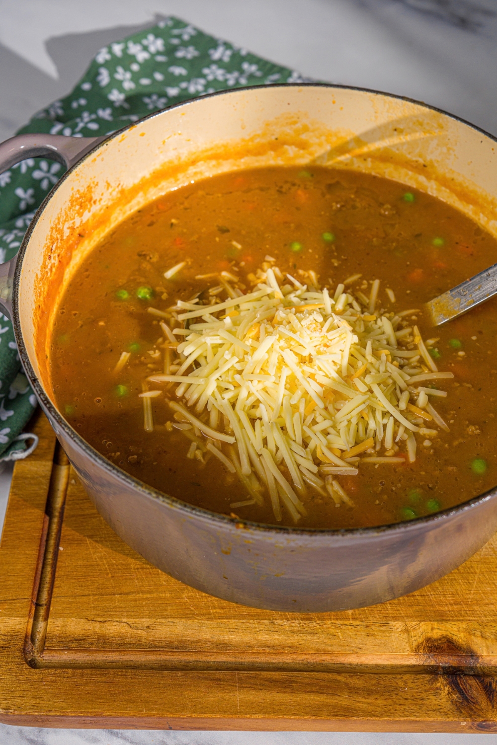 A large pot with shepherd's pie soup topped with shredded cheese. The pot is on a wooden board with a green cloth napkin.