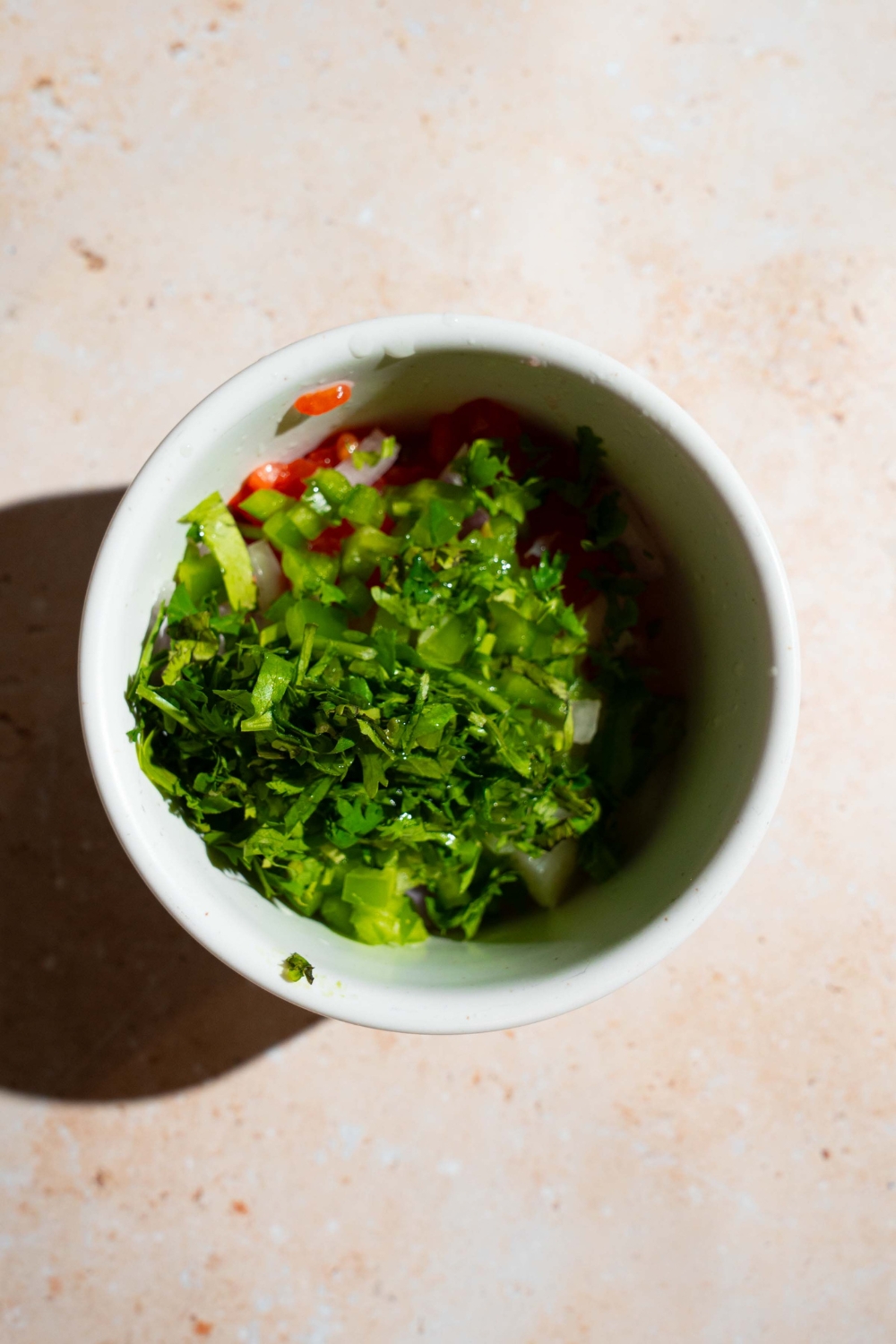 A small bowl with ingredients to make pico de gallo including diced tomatoes, onion, and cilantro. The bowl is on a tan counter.