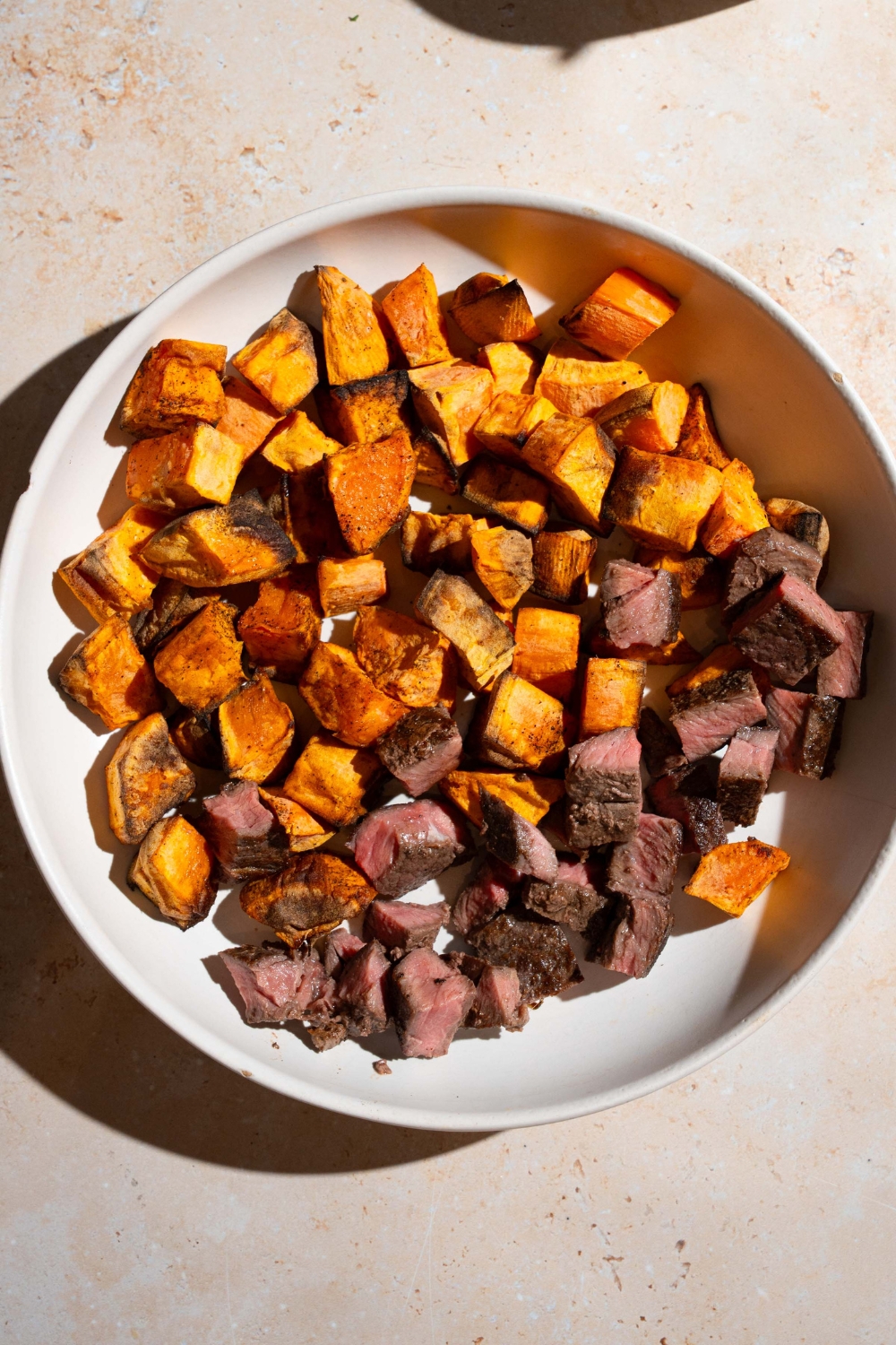 A bowl with cubed fried sweet potatoes and cubed seasoned steak. The bowl is on a tan counter.