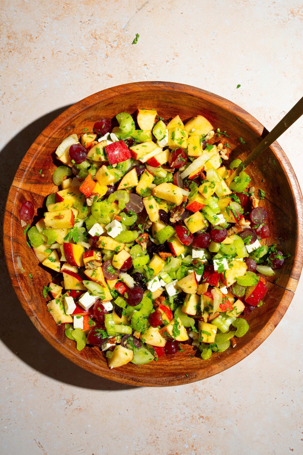 A wooden bowl with apple crunch salad including apples, celery, pecans, feta cheese, parsley, and grapes tossed in a maple dijon dressing. There is a fork in the bowl. The bowl is on a tan counter.