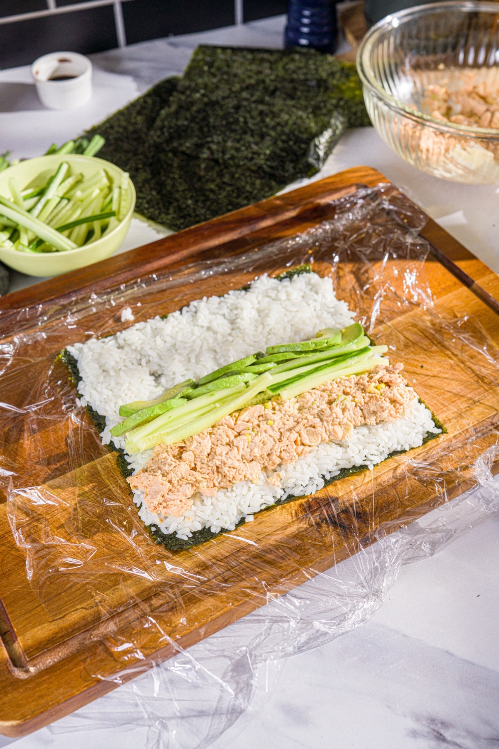 A wooden board lined with saran wrap topped with a seaweed wrap with sushi rice, cucumber, tuna mixture, and avocado.