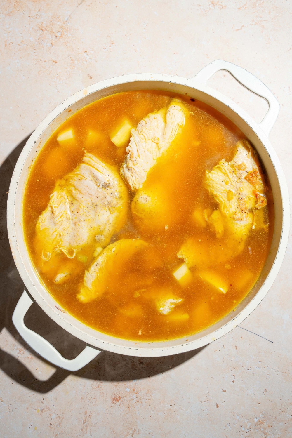A large pot with seared chicken with stock and potatoes. The pot is on a tan counter.