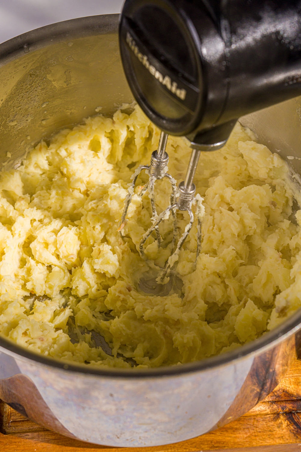 A hand mixer mixing a pot of cooked potatoes. The pot is on a wooden board.